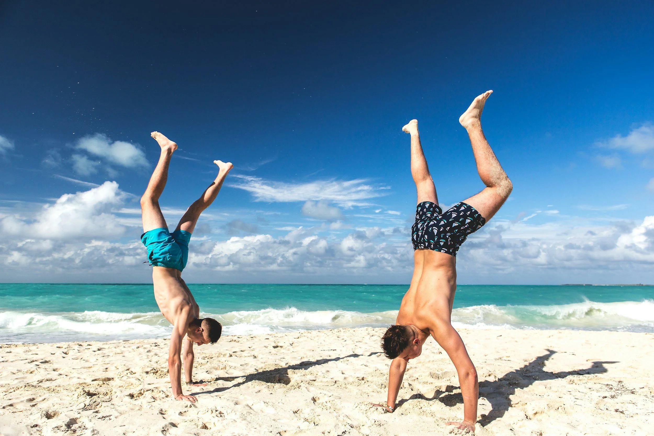 Two boys performing handstands on a sandy beach with ocean waves and cloudy sky in the background.