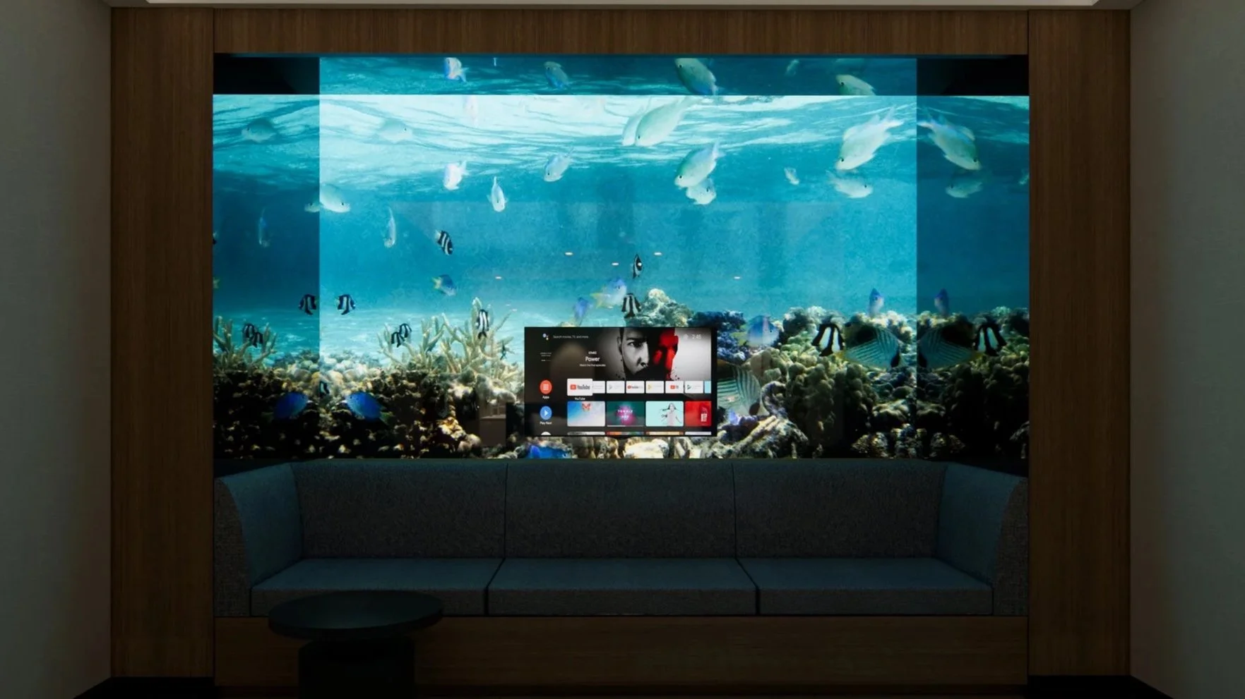 An indoor seating area with a large aquarium wall containing colorful fish, corals, and underwater scenery, with a television screen displaying various app icons and a black and white image of a person's face.