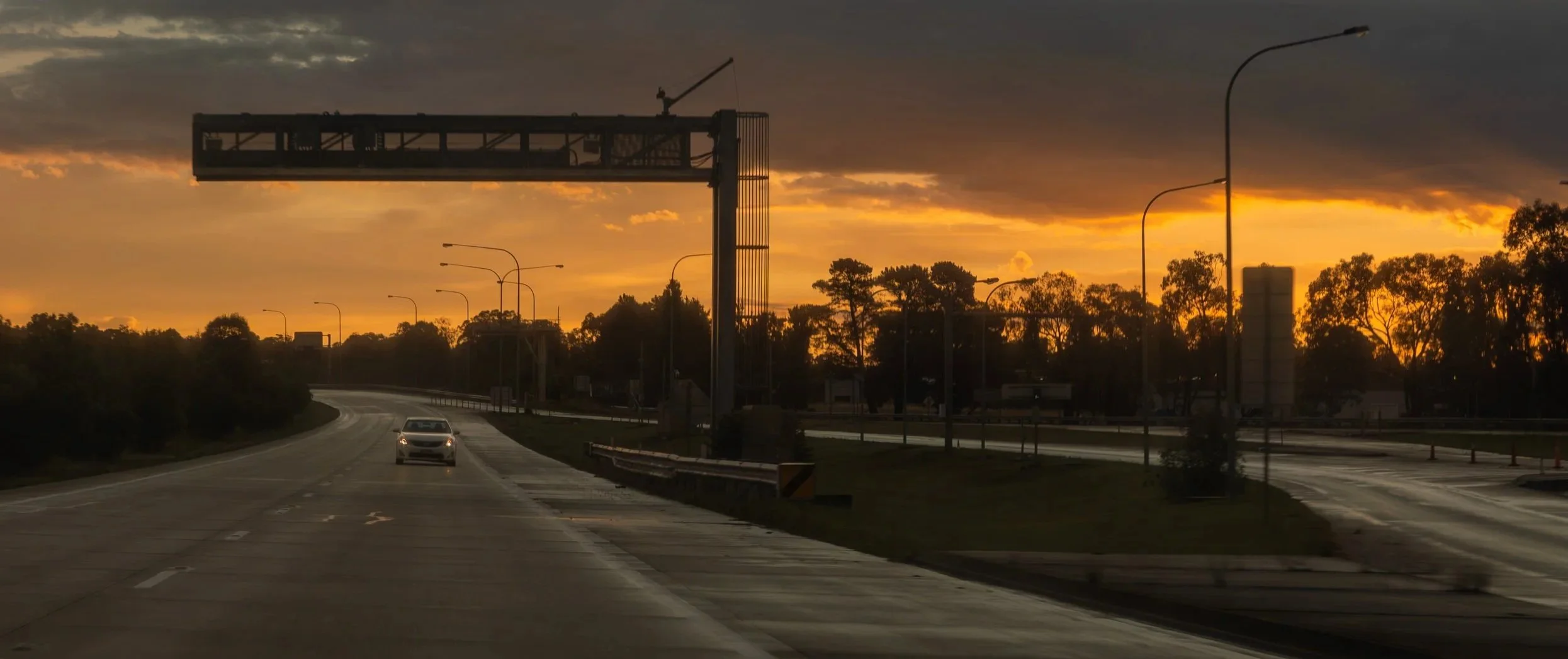 A highway at sunset with a car driving, streetlights, trees, and a large overhead gantry.