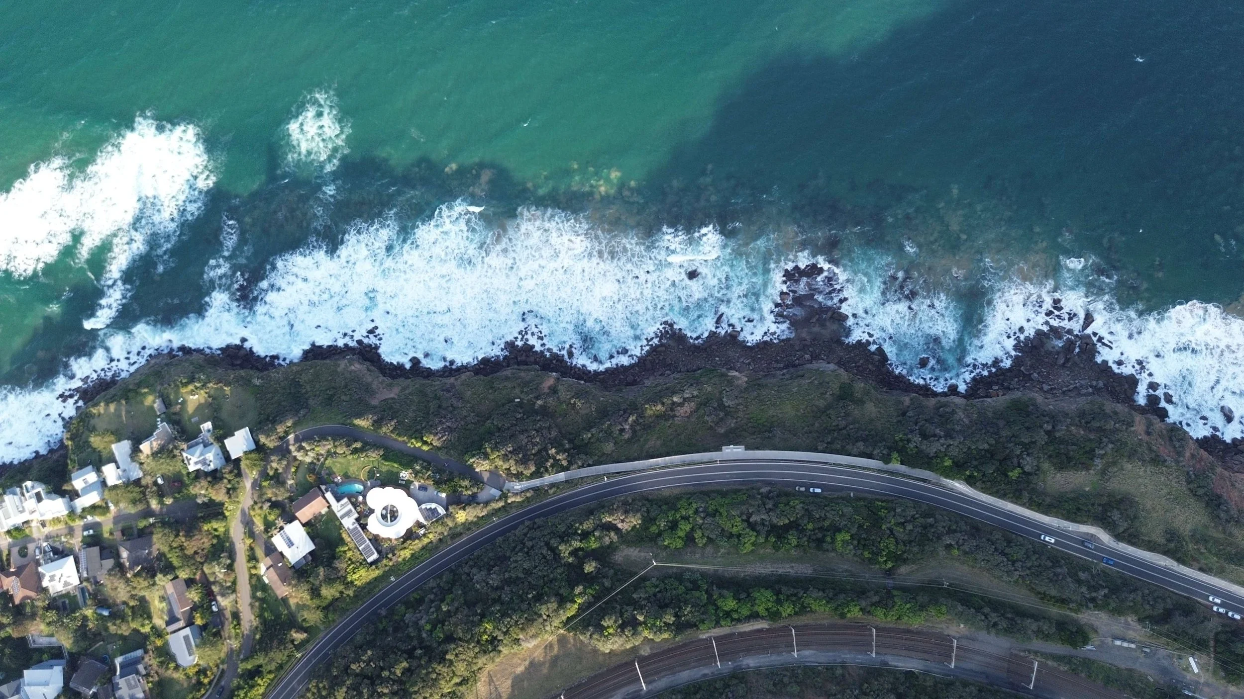 An aerial view of a coastal area showing a road curving along the edge of the land with houses and greenery, and a rocky shoreline with ocean waves crashing against it.