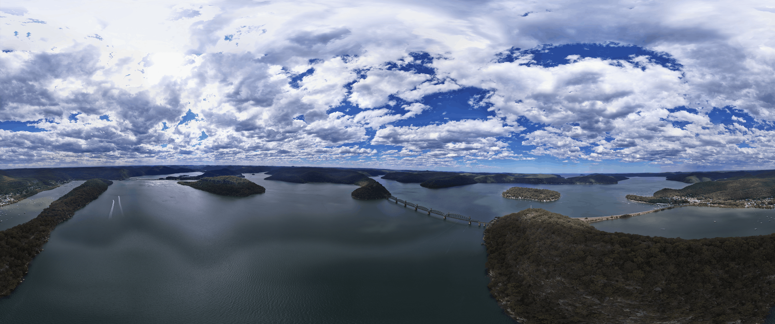 A panoramic aerial view of a large lake with multiple islands and a bridge connecting two sides, surrounded by hilly terrain and a partly cloudy sky.