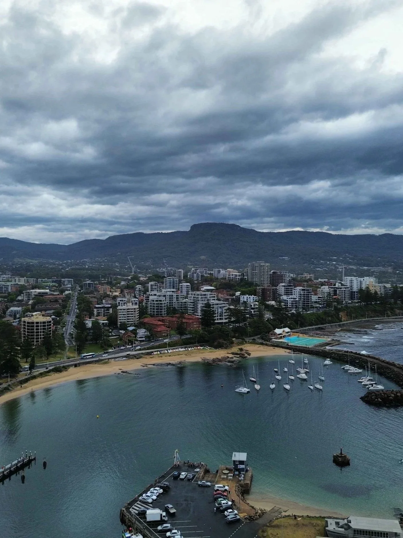 A coastal city with high-rise buildings and a bay with sailboats docked, a sandy beach, and a mountain range in the background under a cloudy sky.