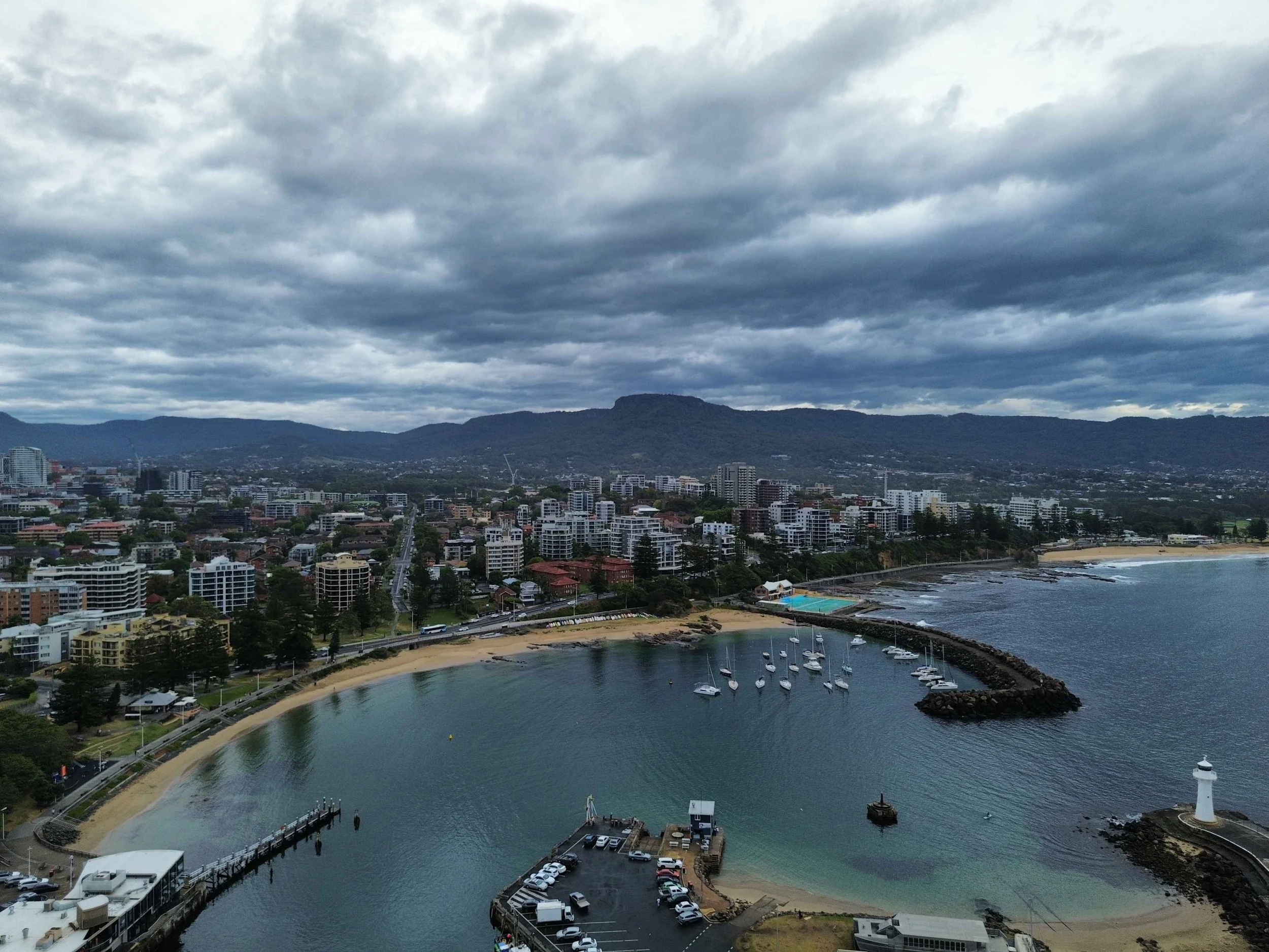 A coastal city with a harbor, sailboats, a lighthouse, and high-rise buildings against a backdrop of mountains and cloudy skies.