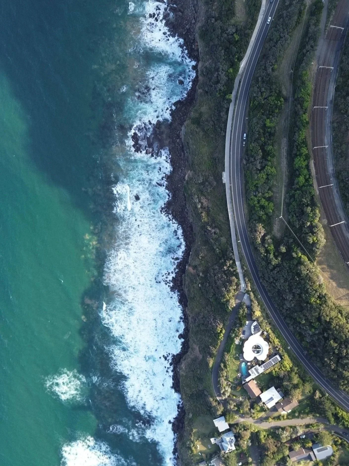 An aerial view of a coastline with waves crashing against rocks, a curving road along the edge, and residential buildings surrounded by trees.