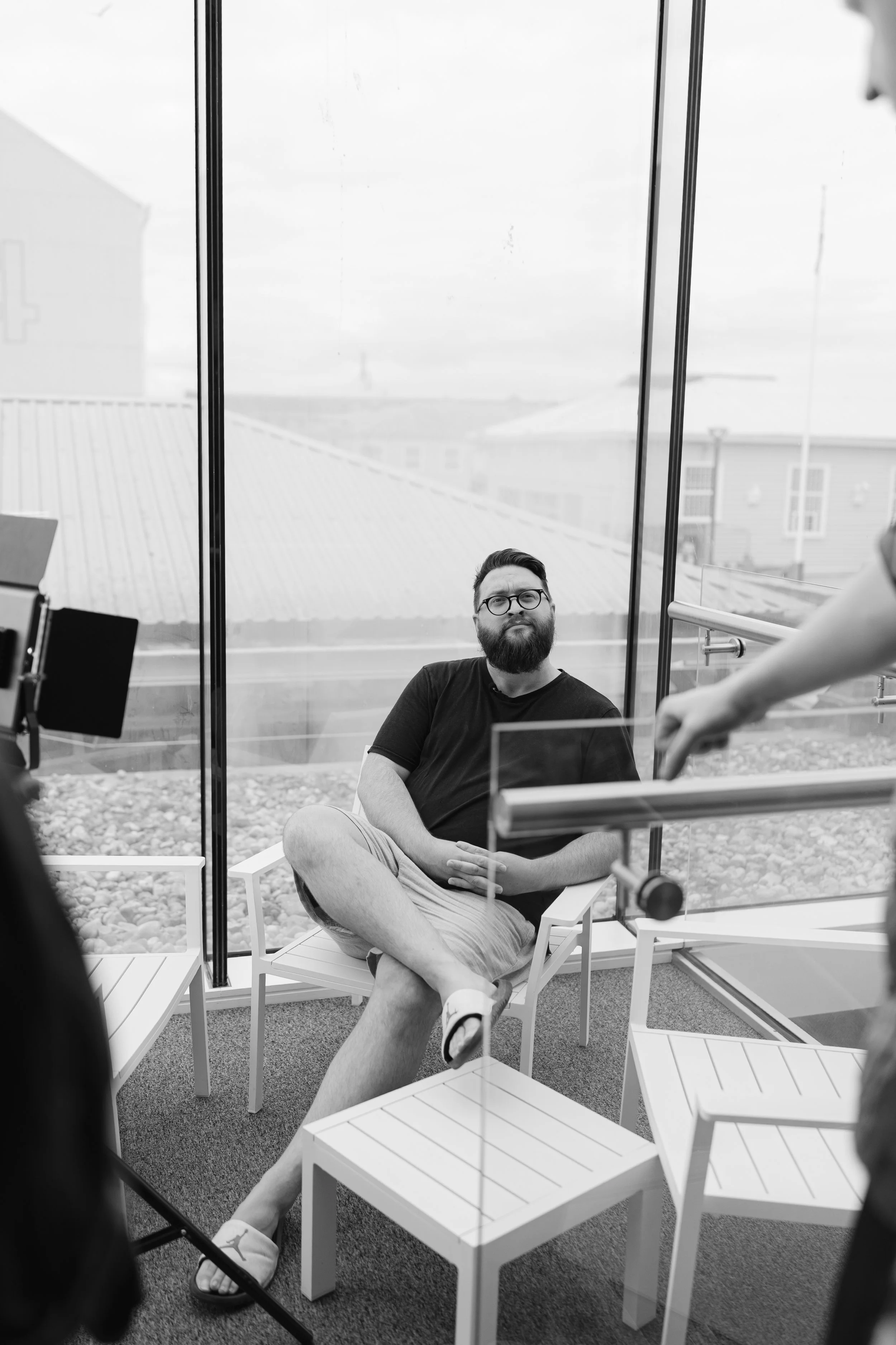 A man with glasses and a beard sitting on a chair with one leg crossed over the other, inside a glass-enclosed space, possibly a studio or interview setup. He is smiling and wearing a black shirt and shorts, with a background of a roof and buildings visible through the glass.
