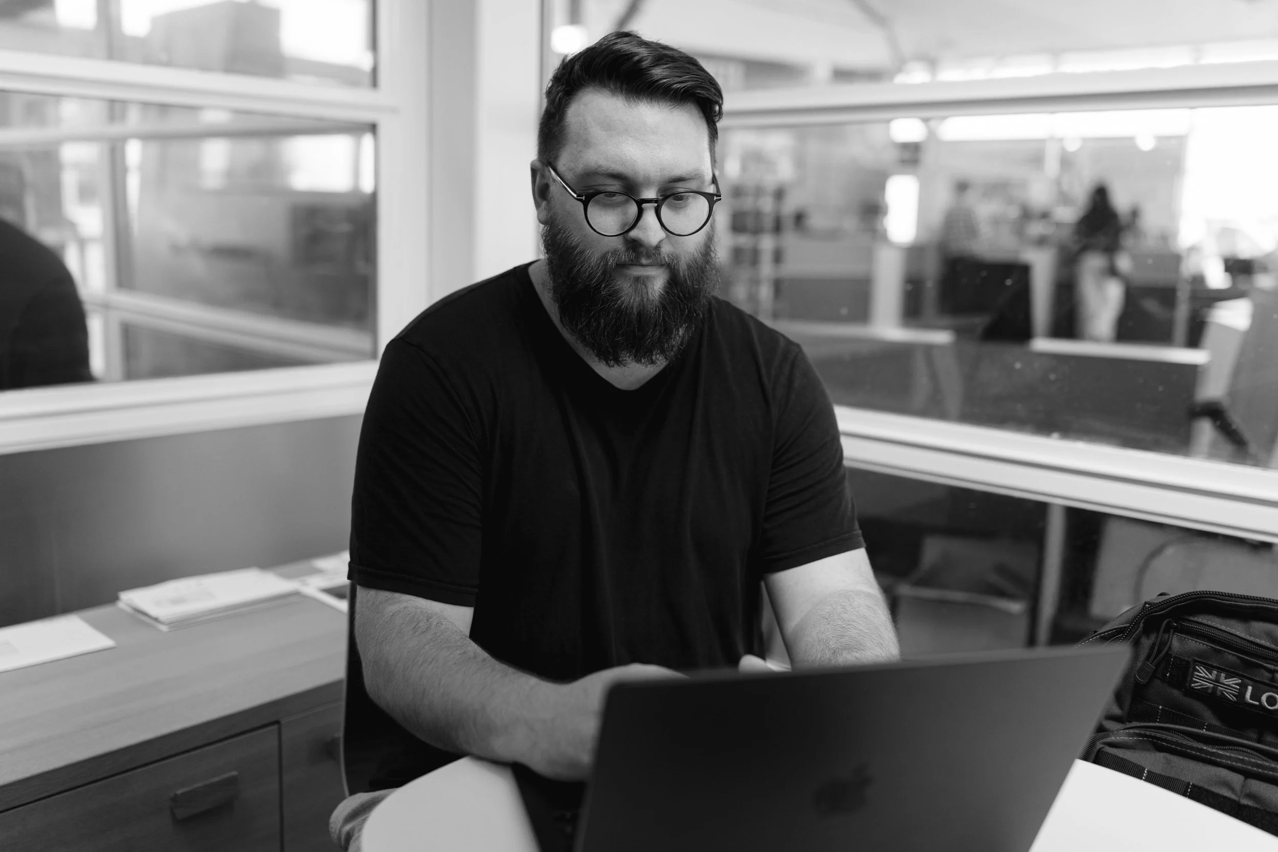 A man with a beard and glasses working on a laptop in an office setting