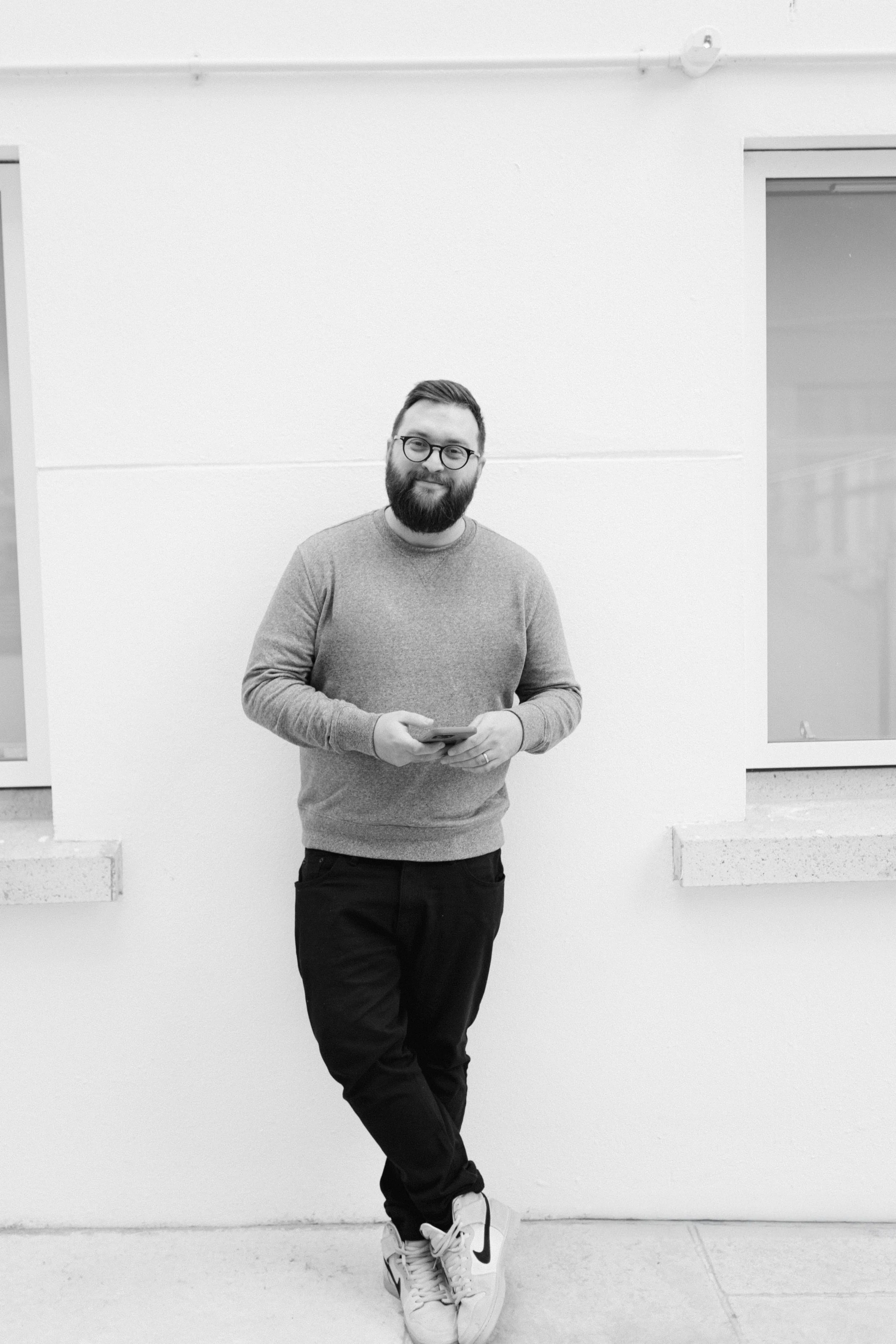 A man with glasses and a beard standing against a plain wall, holding a smartphone, wearing a sweater, black pants, and sneakers.