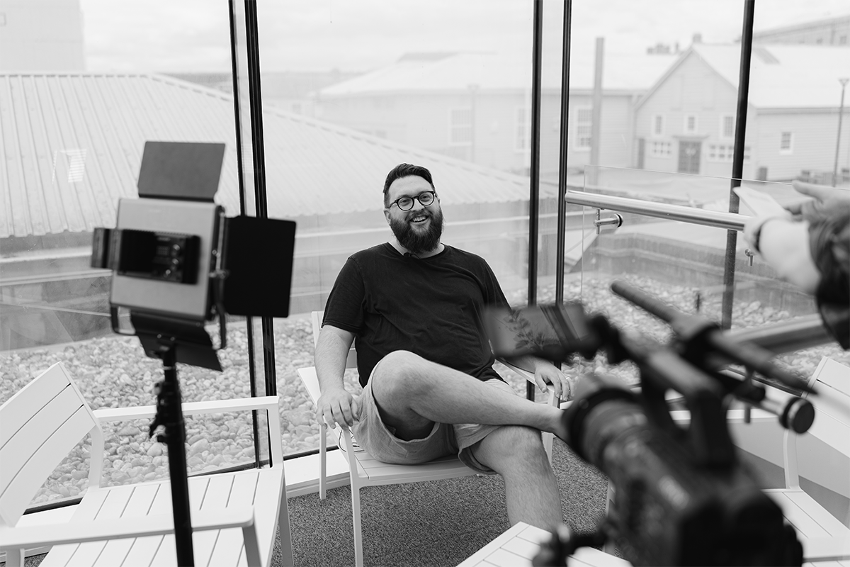 A man with glasses, a beard, and a black t-shirt sitting on a bench inside a glass-walled room, smiling, with a camera and a person with a tablet nearby.