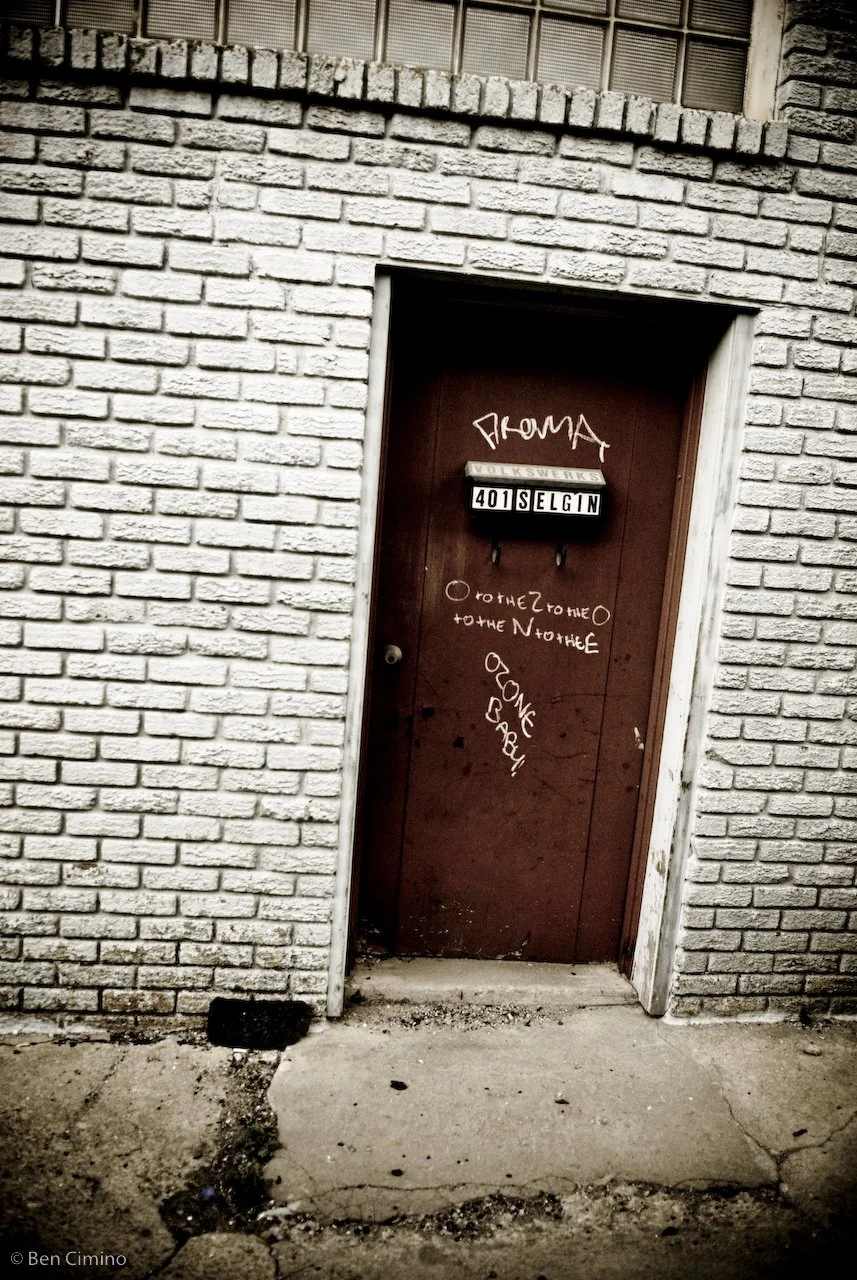 A brown door set into a white brick wall with graffiti and signs, including a sign reading '401 Selgin' and handwritten messages in white chalk, next to a concrete sidewalk. The door has a small mail slot and graffiti text.