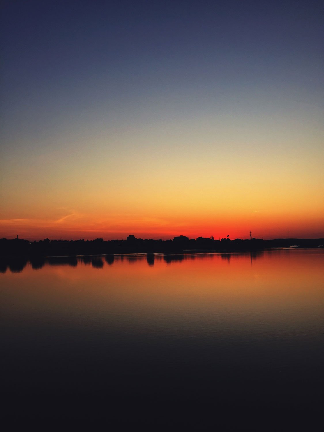 Sunset over a body of water with a colorful sky and dark silhouette of trees and buildings in the distance.