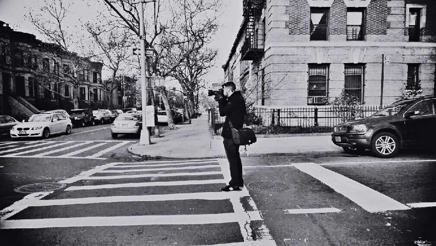 Black and white photo of a man waiting to cross the street at a crosswalk, holding a camera with a backpack at his side. The street is lined with cars and old buildings with leafless trees in the background.
