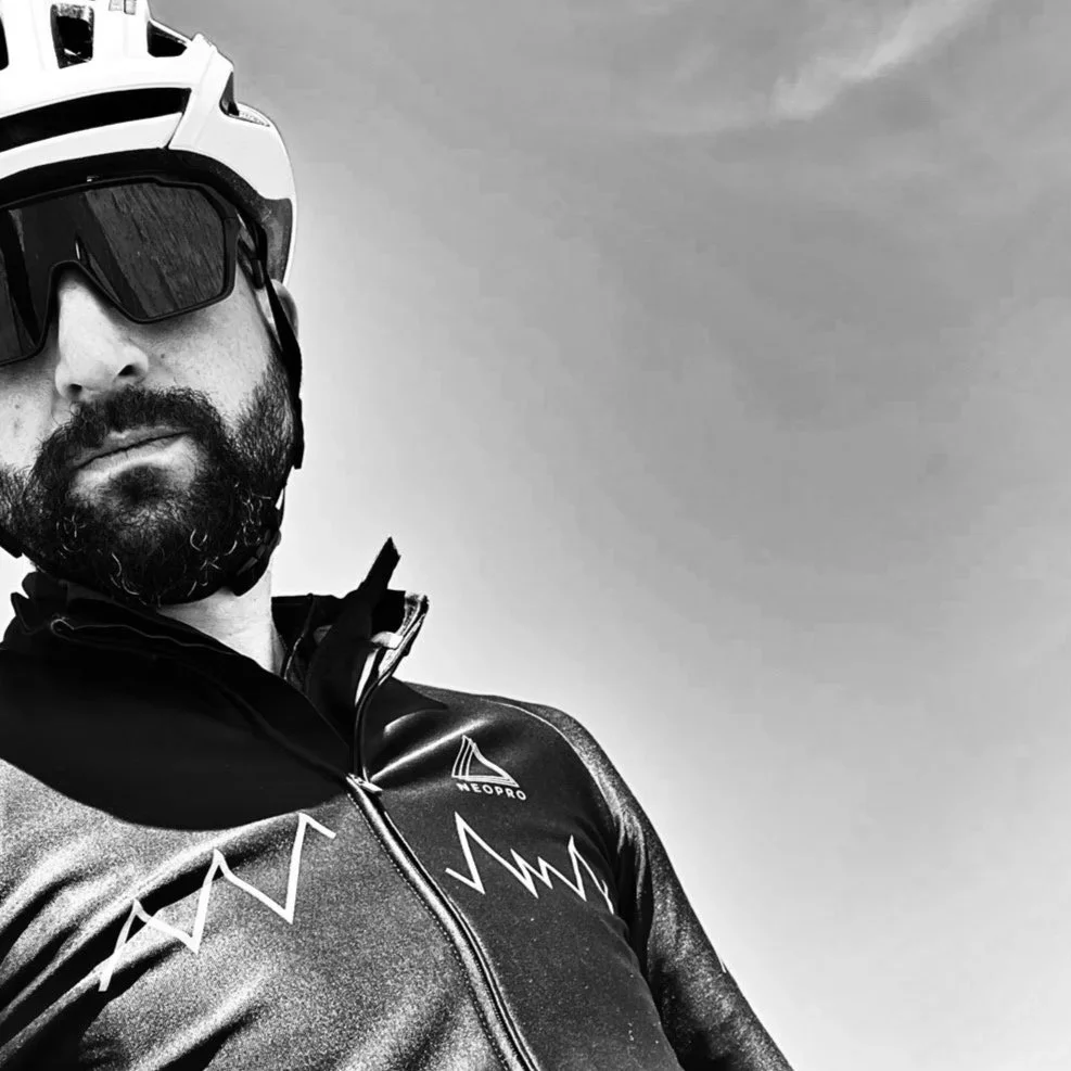 A man wearing a cycling helmet and sunglasses, dressed in a cycling jersey with mountain and heartbeat graphics, outdoors under a cloudy sky.