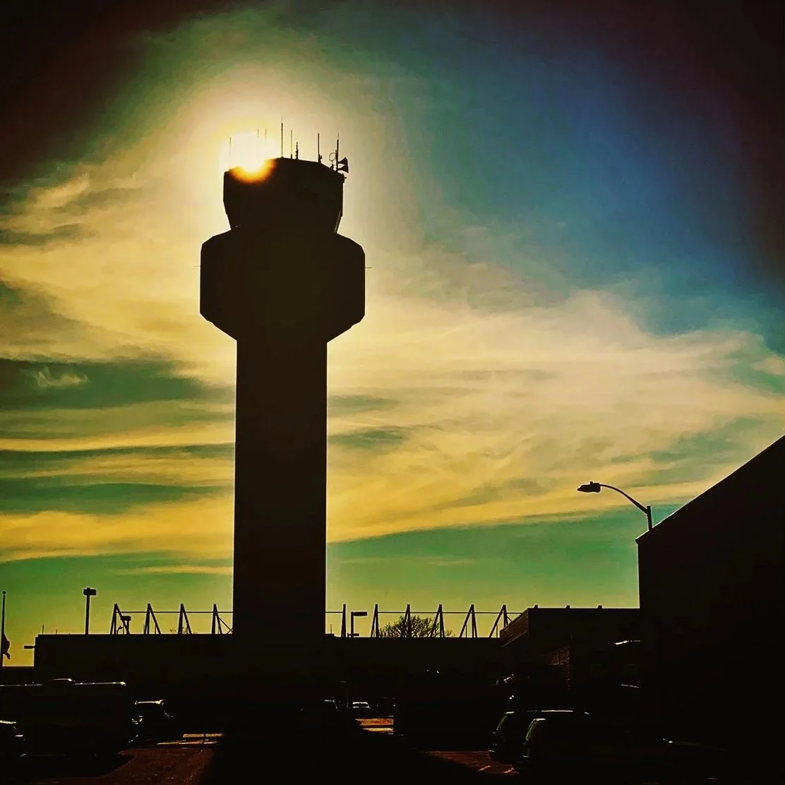A dark silhouette of an airport control tower with the sun setting or rising behind it, casting a glow around its top. The sky has streaks of clouds with a gradient of colors from green to yellow and blue. The foreground shows parked cars and a streetlamp.