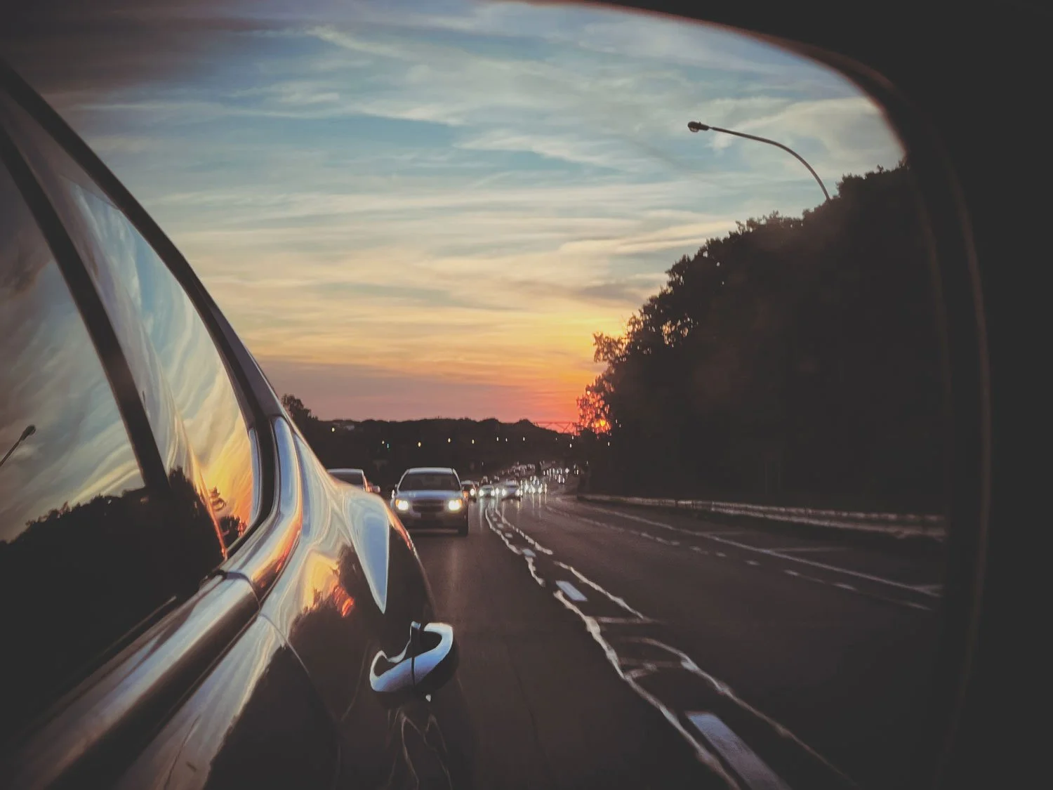 View of a car on a highway during sunset, seen through the side mirror, with other cars in the distance and trees on the side of the road.