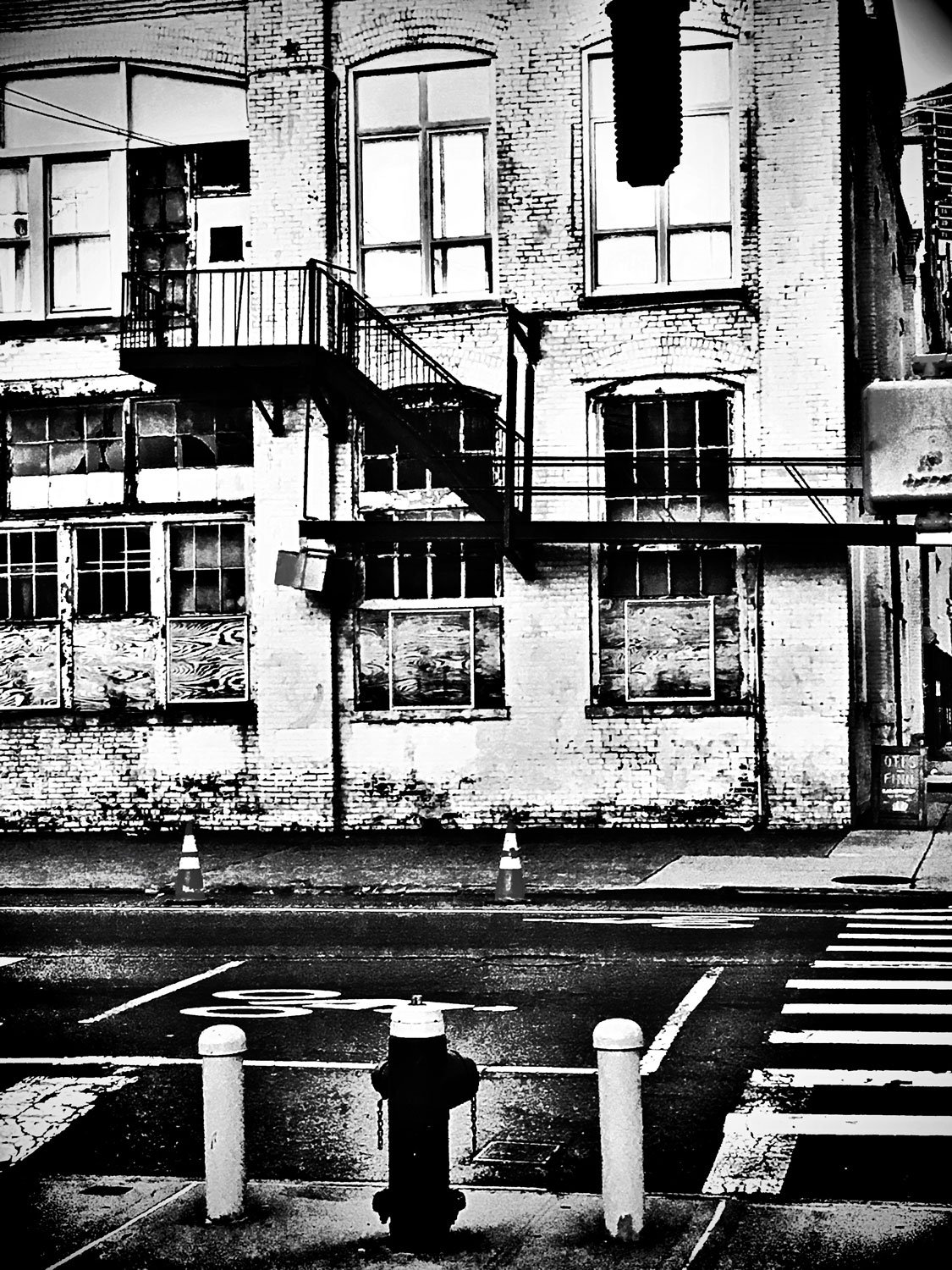 Black and white photo of a brick building with multiple windows, some boarded up. A fire escape staircase runs down the front of the building. In the foreground, a wet street with traffic cones, a fire hydrant, and a crosswalk.