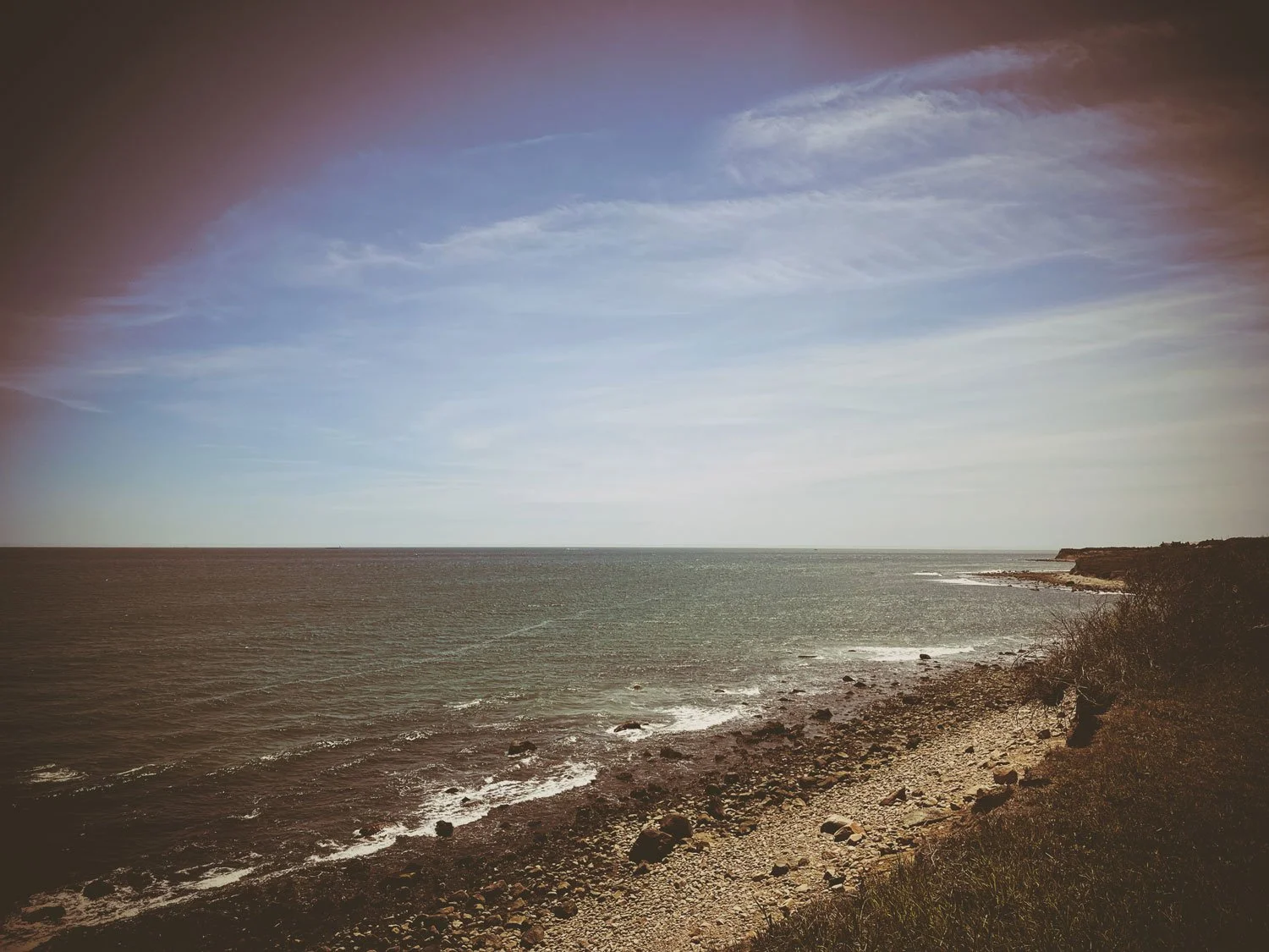 A rocky shoreline along a large body of water with a partly cloudy sky overhead.