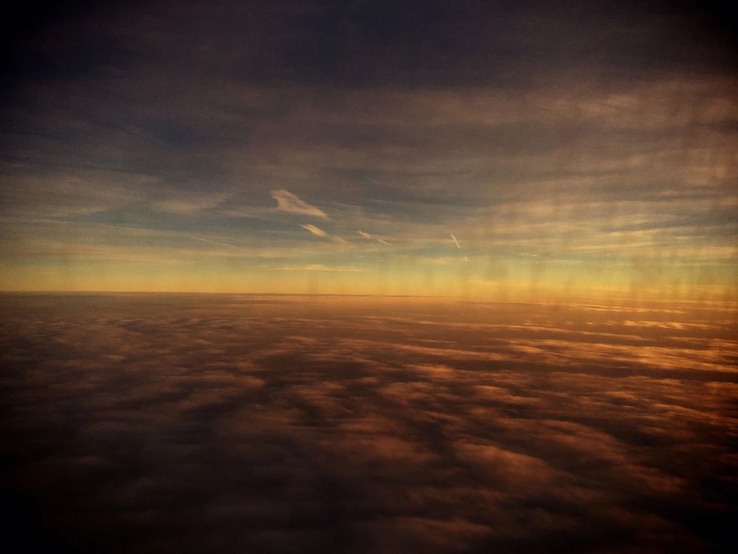 Sky viewed from an airplane window during sunset, with clouds below and a colorful horizon.