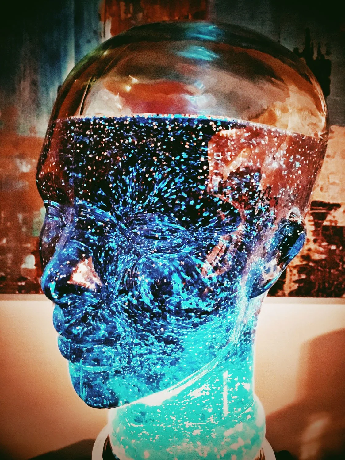 A glass sculpture of a human head illuminated with blue and red lights, creating a cosmic, galaxy-like effect with swirling patterns inside.
