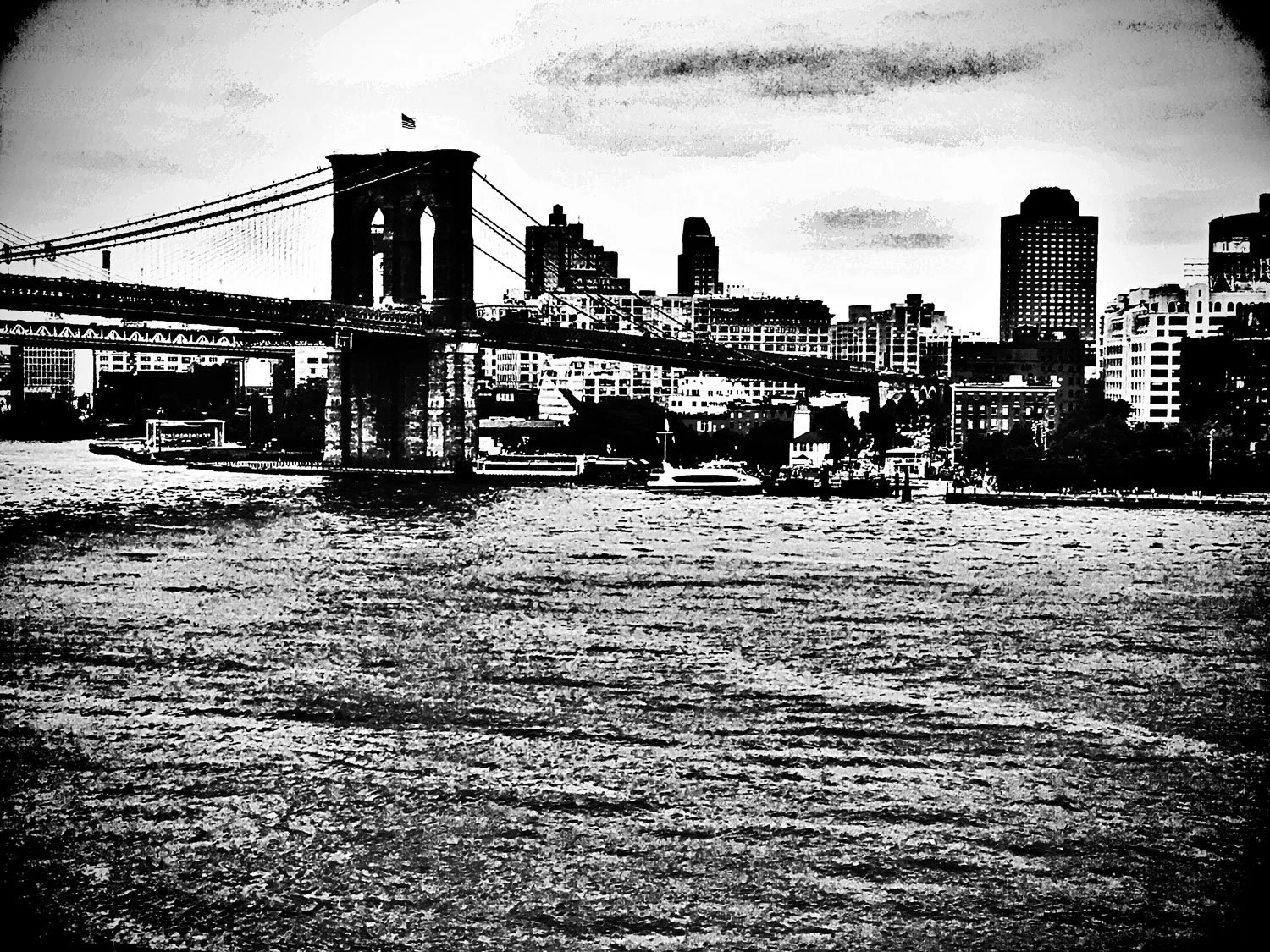 Black and white photo of the Brooklyn Bridge with the Manhattan skyline in the background, taken from across the river.