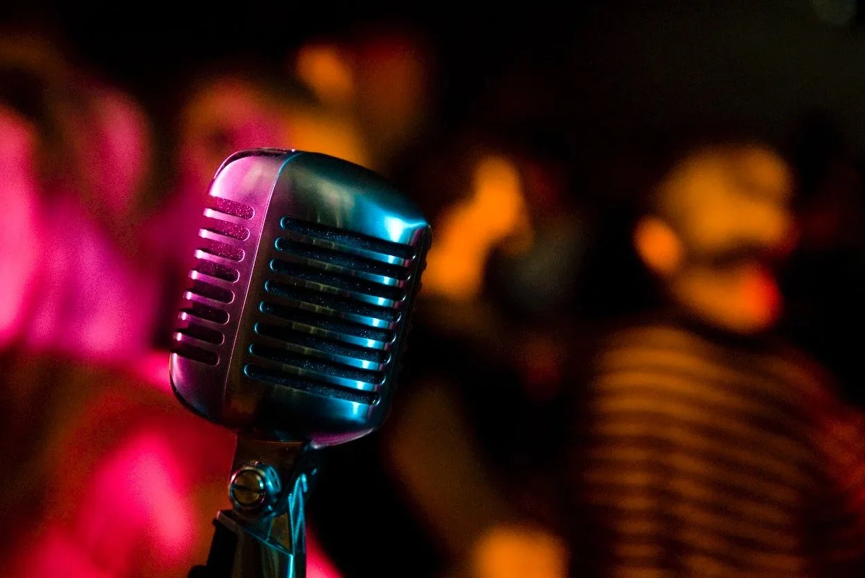Close-up of a vintage microphone with blurred audience members in the background, illuminated with colorful stage lights.