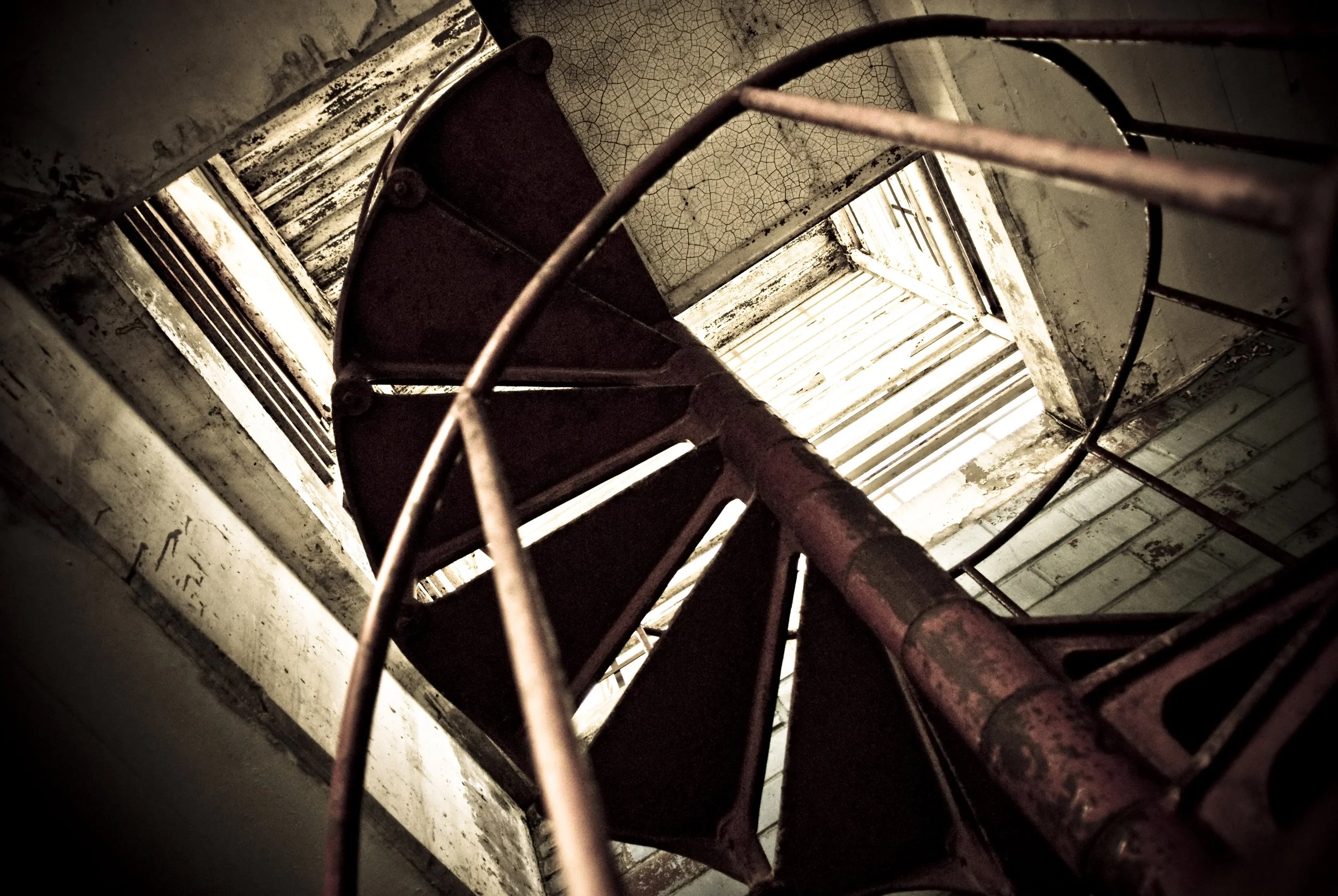 An old, rusty spiral staircase viewed from the top, looking downward, with peeling paint and worn steps.