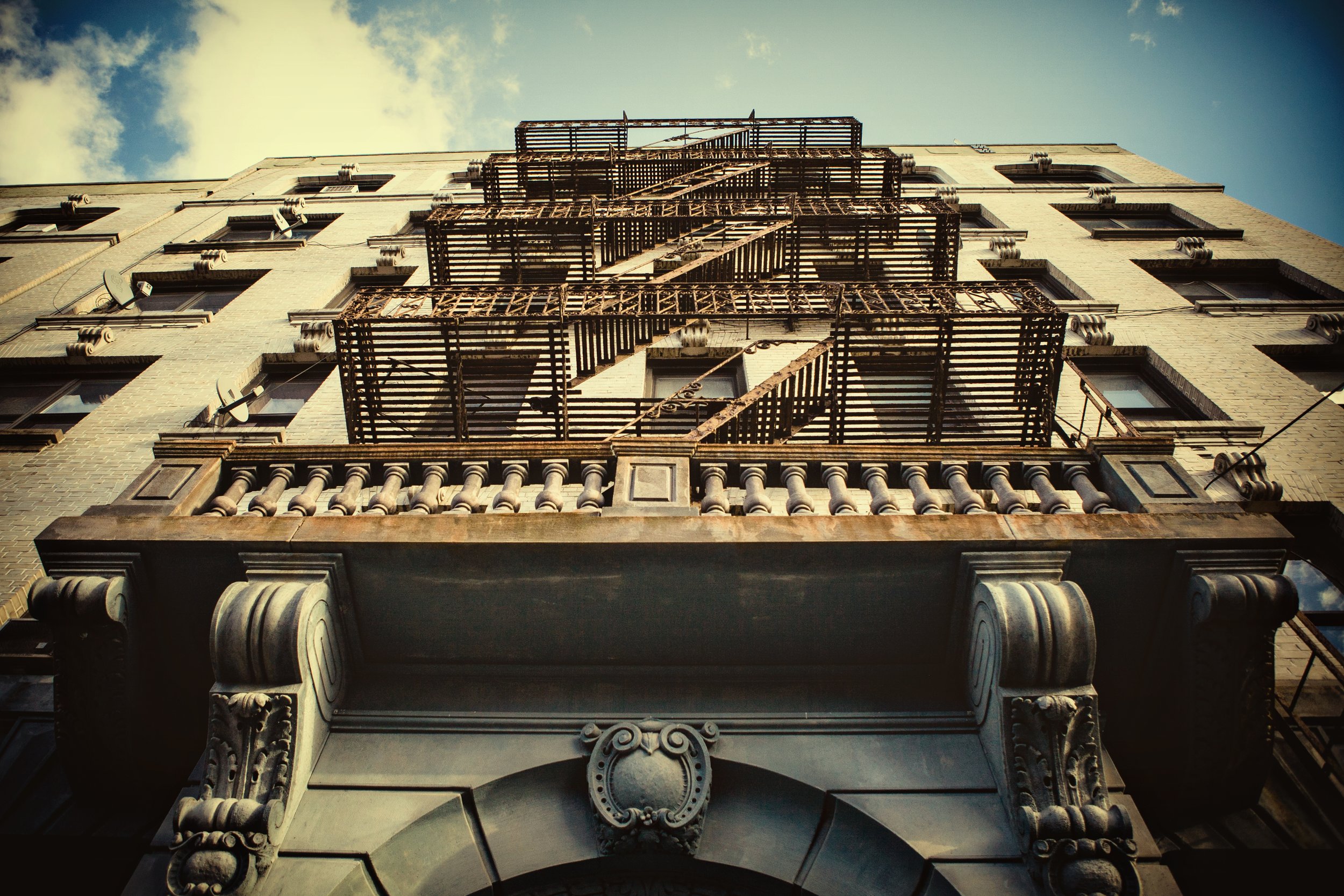 Low-angle view of a building's exterior showing fire escape stairs and windows on a brick facade, with a partly cloudy sky in the background.