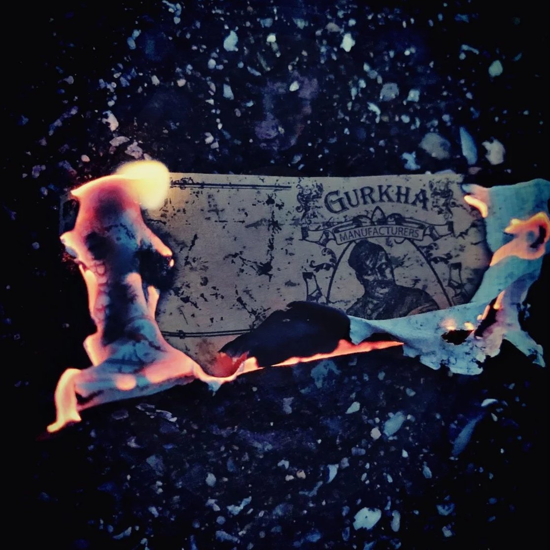 Burnt piece of paper with the word "Gurkha" and a logo, lying on a dark gravel surface, partially burnt with flames around the edges.