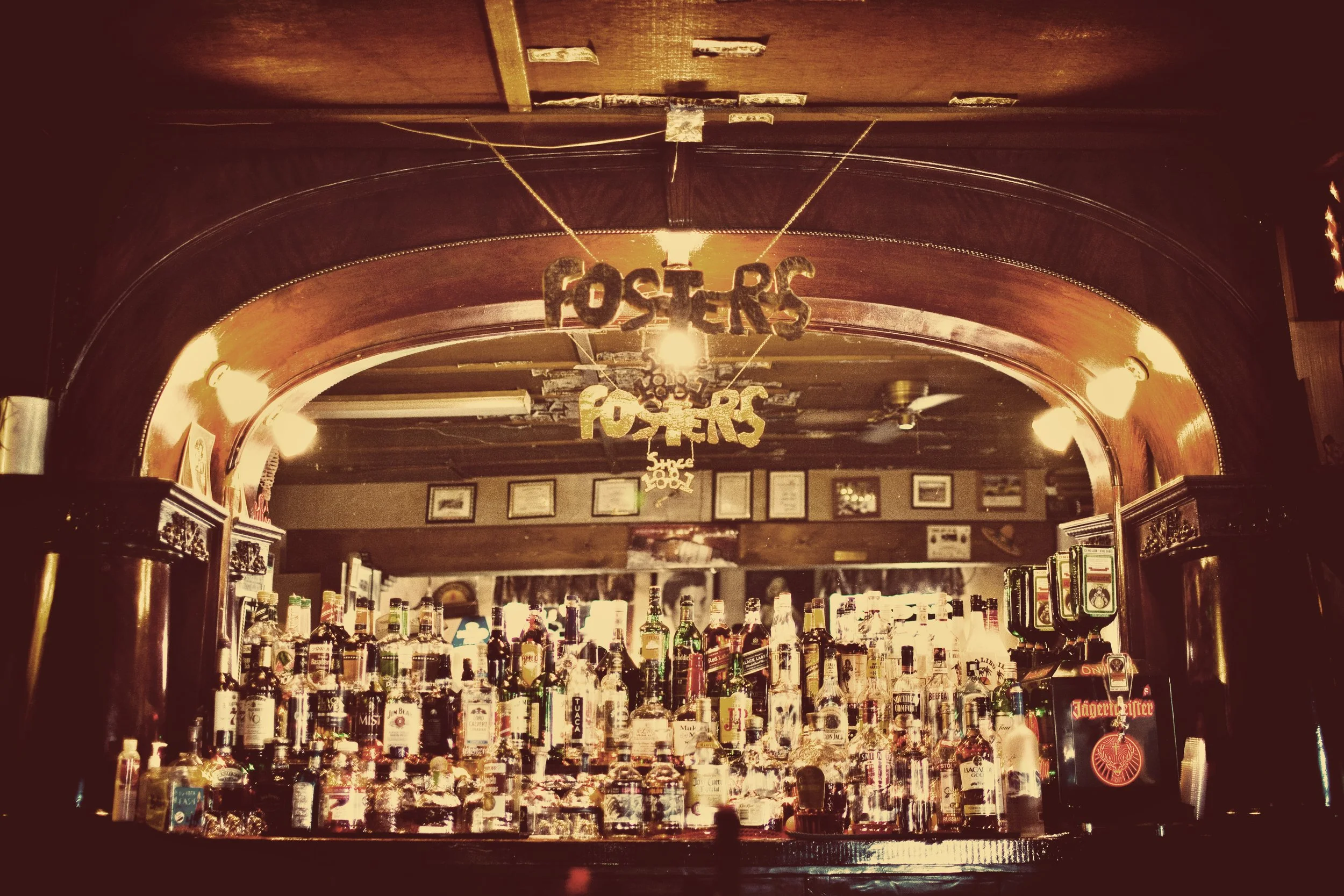 Bar with liquor bottles behind the counter, with a wooden arch above and hanging signs that say 'FOSTERS'.