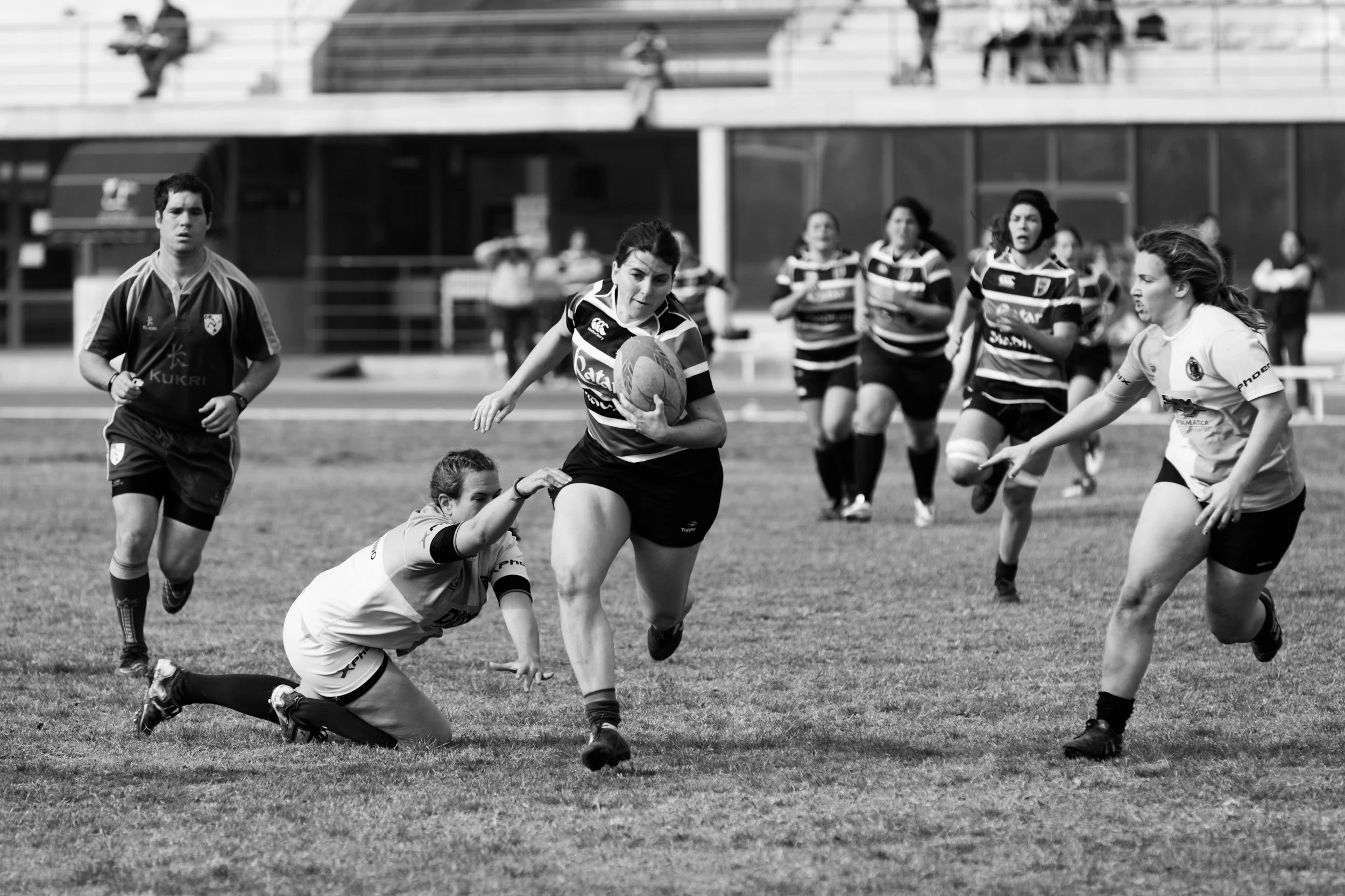 A black and white photo of women's rugby game. Several players are running on the field, with one player in the center holding the rugby ball, while an opponent is on the ground attempting to tackle her. A referee is visible on the left side of the image, and spectators are in the background.