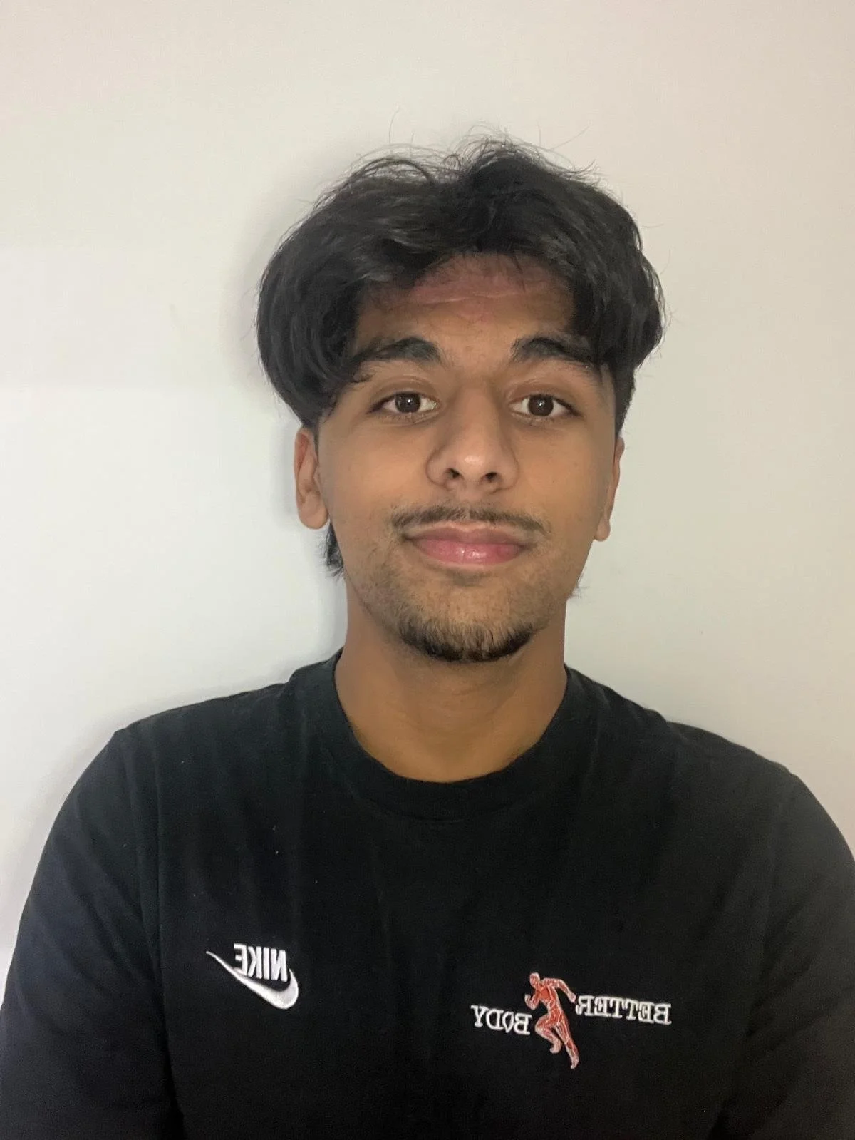 A young man with dark hair and a slight beard, smiling, wearing a black Nike t-shirt with print on the front, standing against a plain white wall.