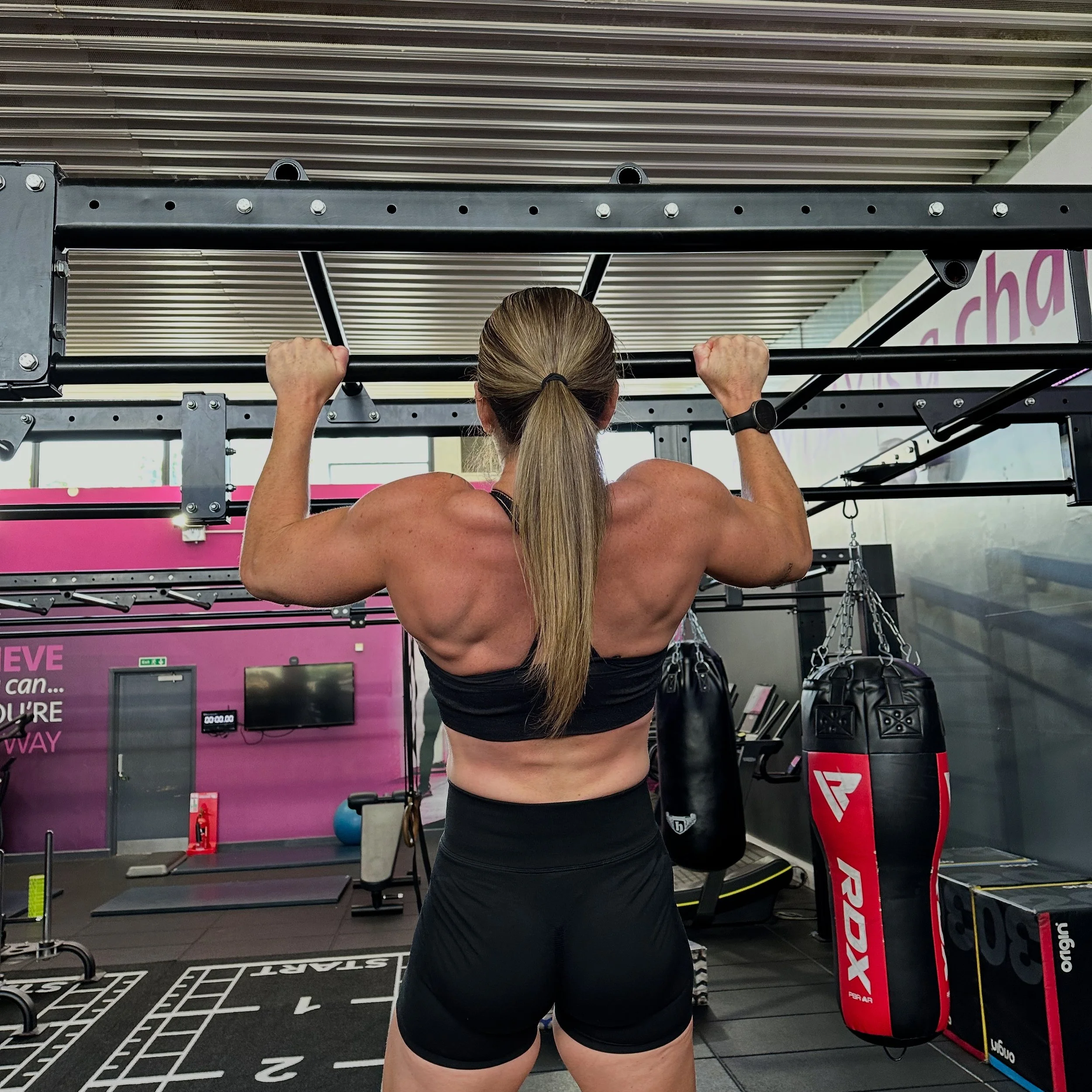 A woman with long blonde hair tied back, wearing a black sports bra and black shorts, is performing pull-ups on a black metal pull-up bar in a gym. The gym has a purple wall with motivational text, and various exercise equipment including a boxing punching bag and a mat area.