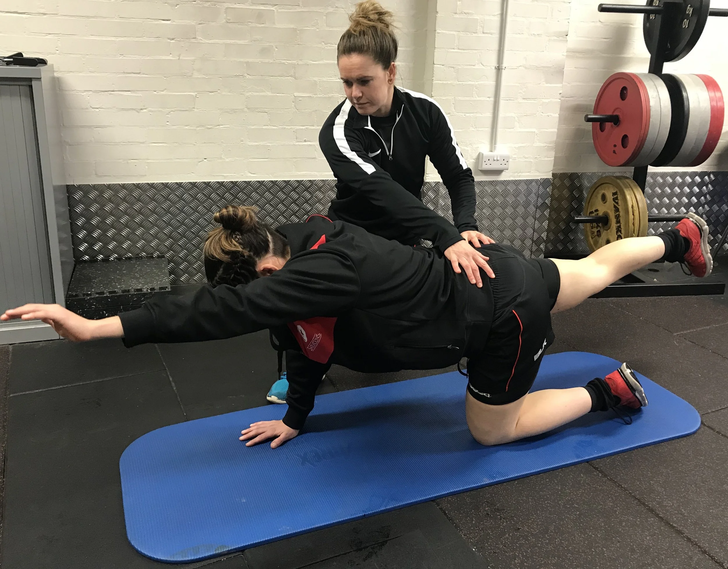 A woman and a trainer are doing a plank exercise on a blue exercise mat in a gym. The woman is in a kneeling position with one arm extended forward, and the trainer is assisting her, placing a hand on her back for support.