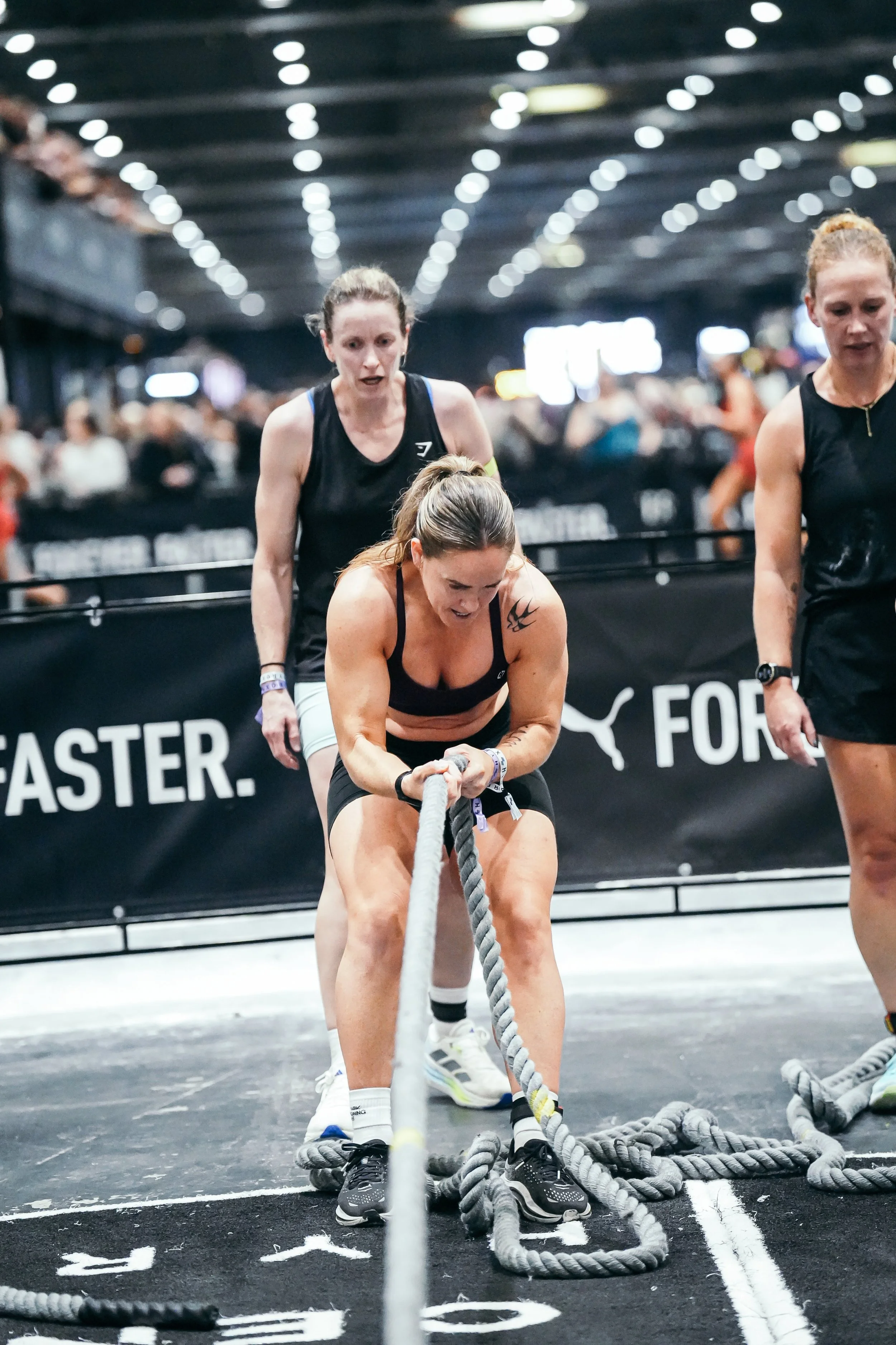 Three women participating in a fitness competition pulling heavy ropes in an indoor gym with blurred spectators in the background.