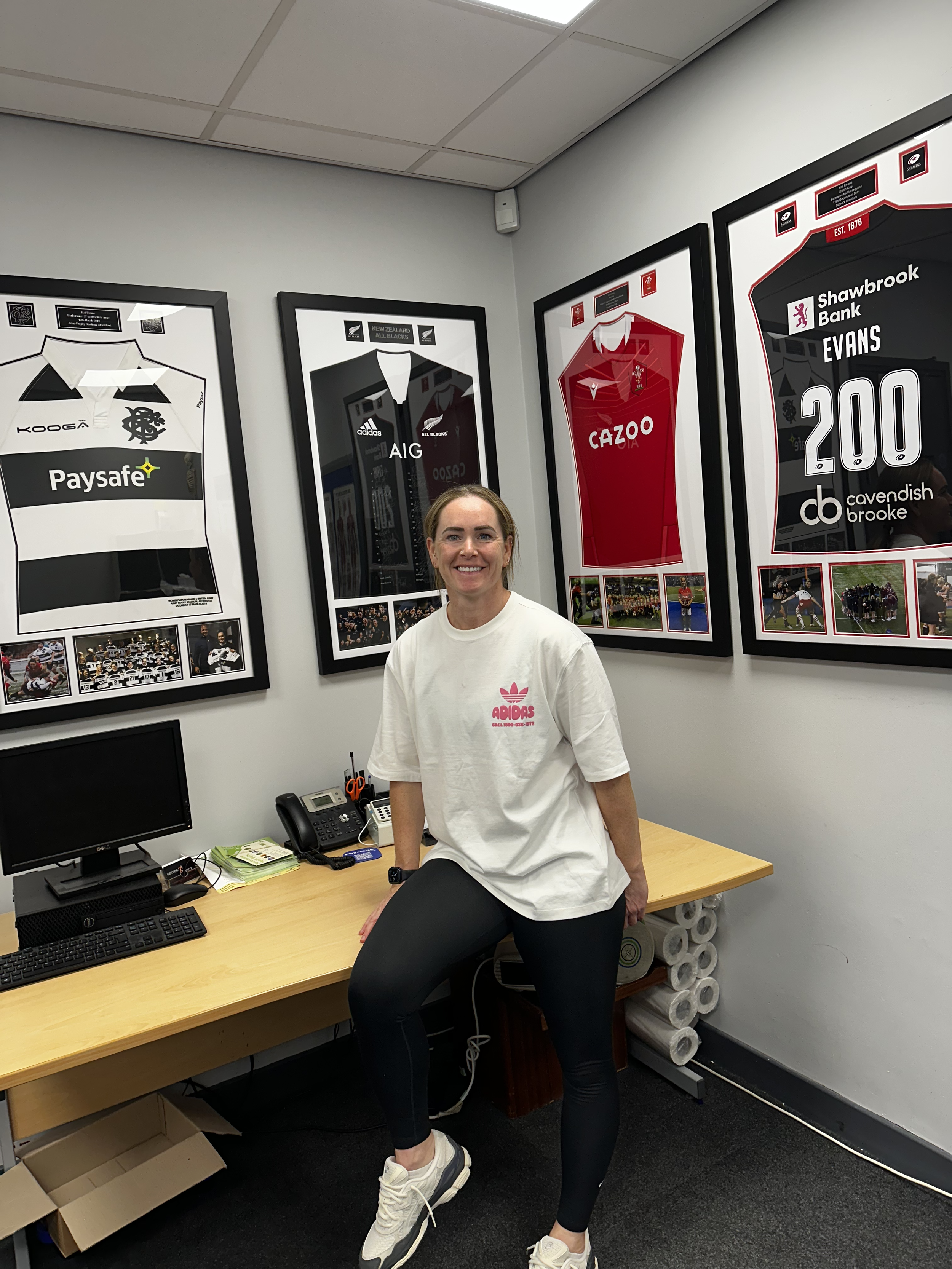 A smiling woman standing in an office, wearing a white Adidas t-shirt, black leggings, and sneakers. She is resting one leg on a chair, with office equipment and framed sports jerseys on the wall behind her.