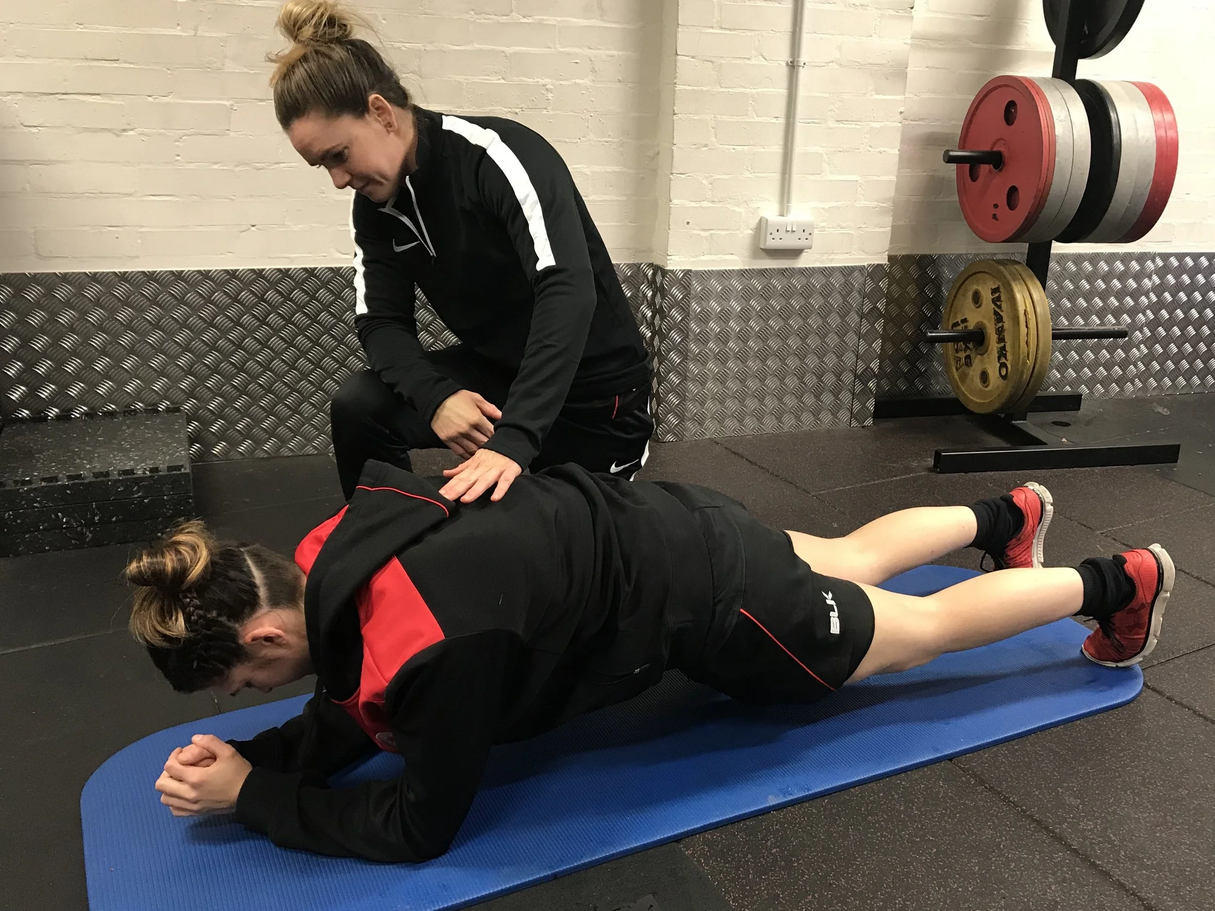 Two women in athletic clothing are performing a plank exercise on a blue yoga mat in a gym. One woman is supporting the other, who is lying face down with elbows bent and head down. The gym has a white brick wall and weight plates on a rack in the background.