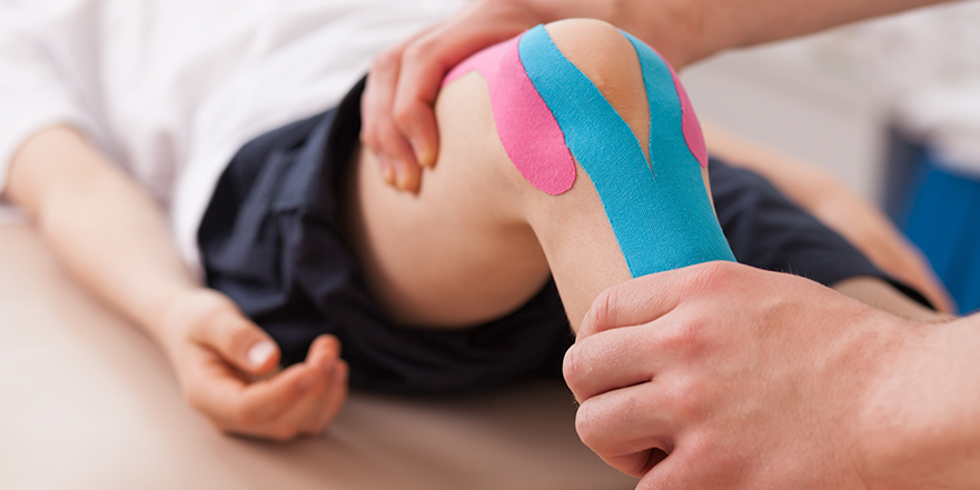 Child lying on a table receiving a knee injection with colorful athletic tape on the knee.