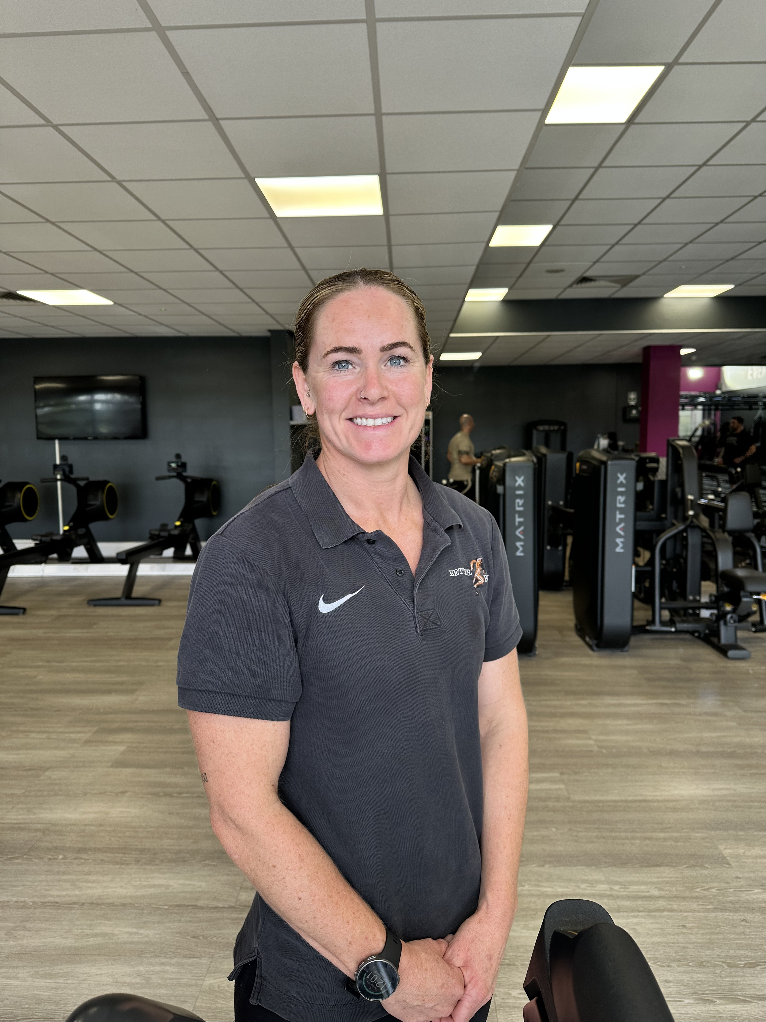 A woman with blue eyes and a nose piercing standing in a gym, smiling, wearing a black polo shirt with a logo, with gym equipment and people working out in the background.