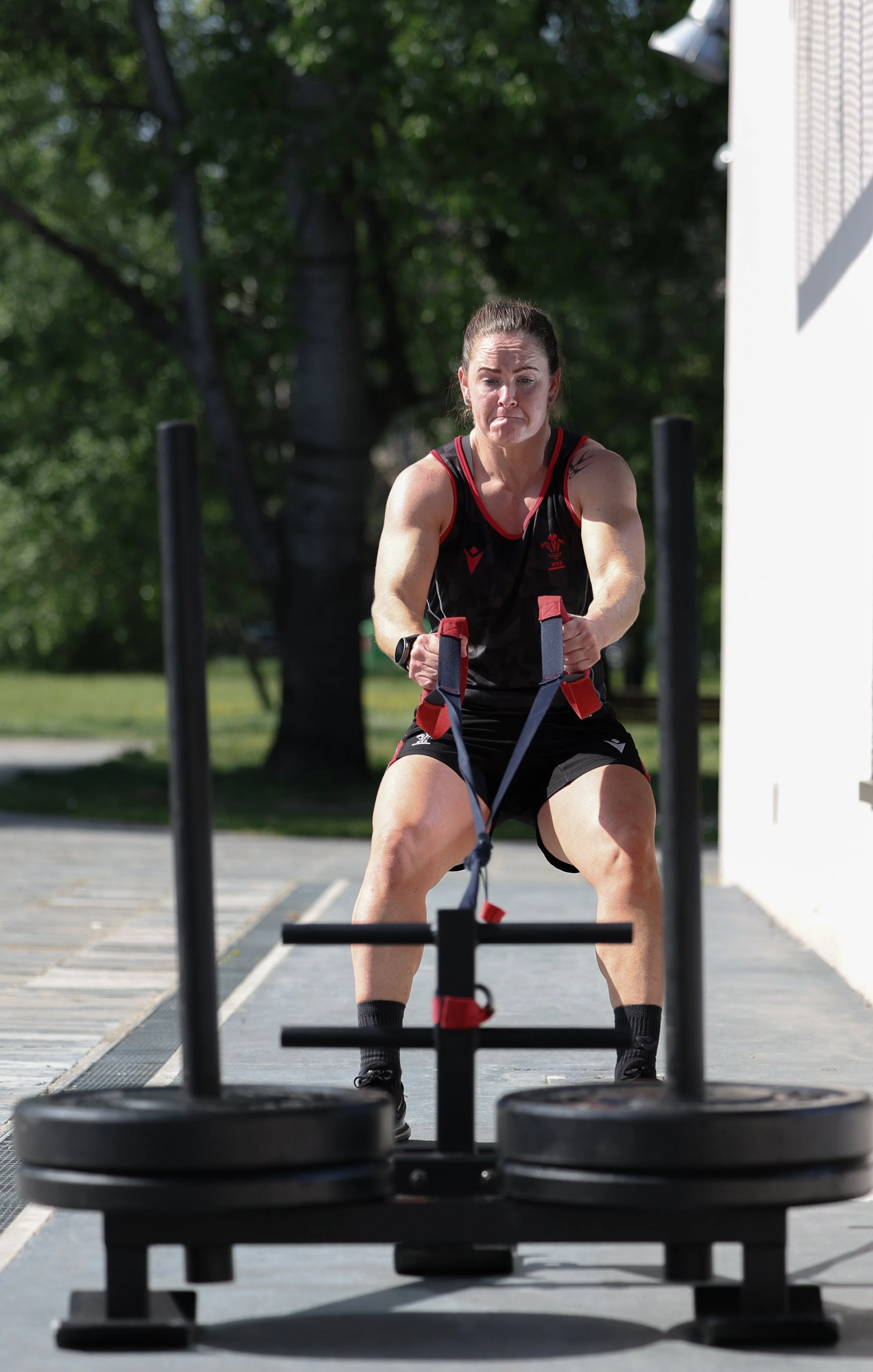 Woman participating in an outdoor workout using a sled push, with green trees and a white building in the background.