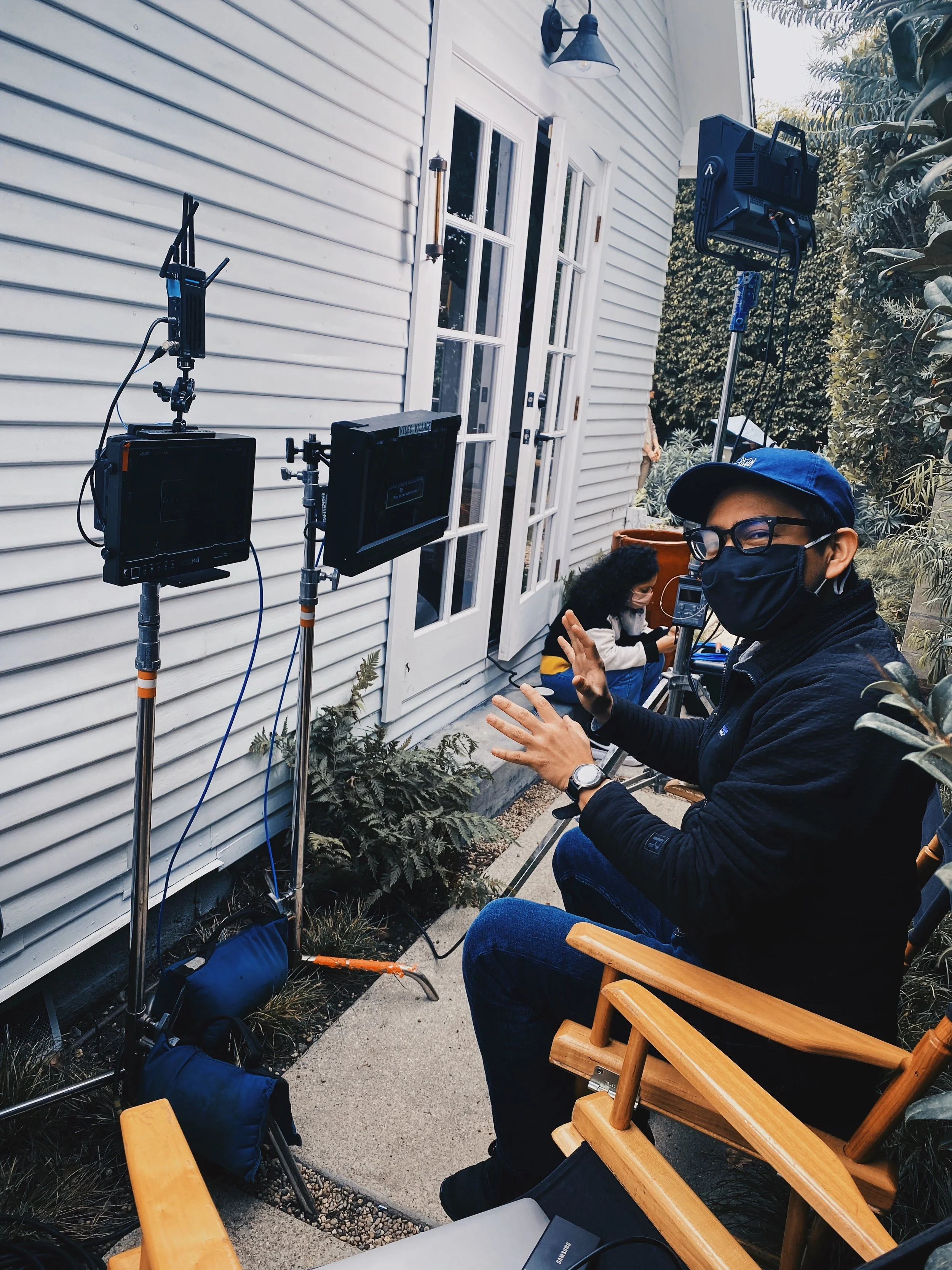 Filming crew setting up equipment outside a white house with double doors and plants nearby.