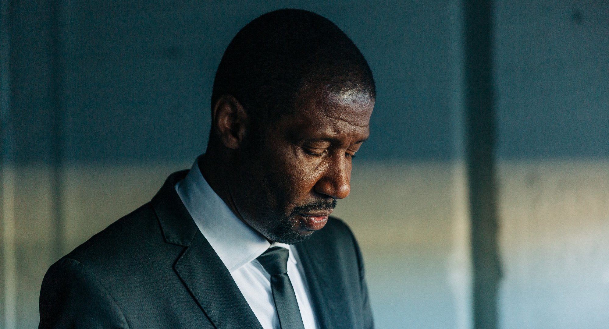 Et bilde av en mann i en svart dress og hvit skjorte med sort slips, som ser nedover med et alvorlig uttrykk.