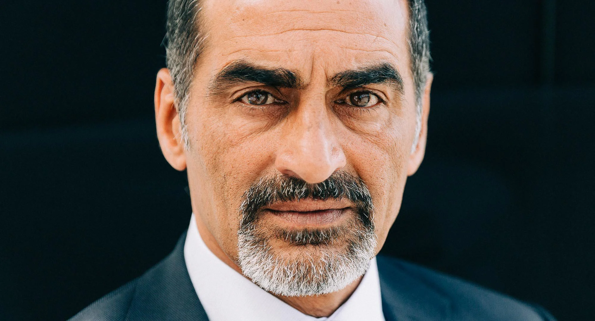 Nærbilde av en mann i dress med grått skjegg og mørkt hår, ser direkte på kamera.