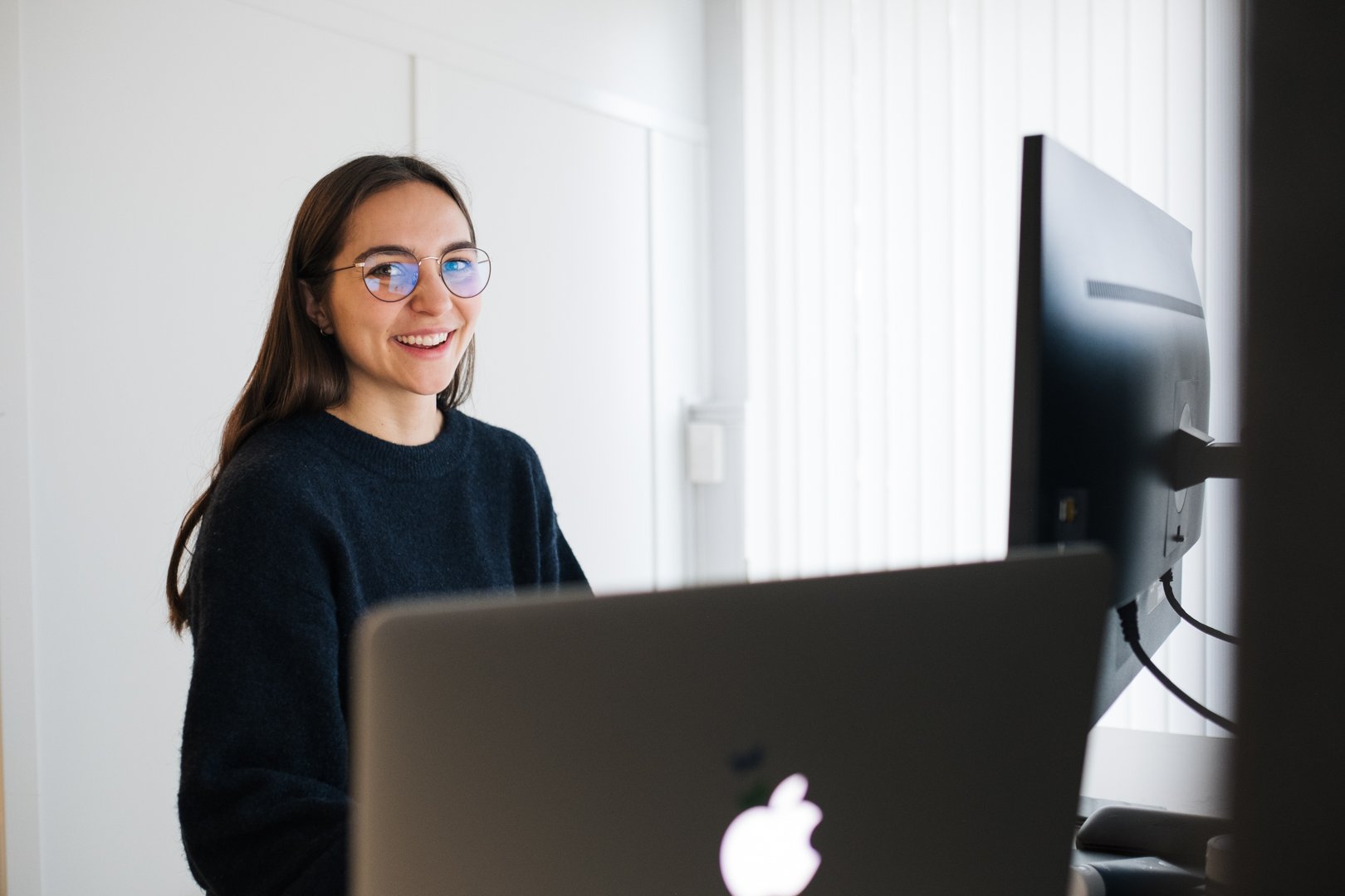 En smilende kvinne med briller sitter foran en datamaskin i et lyst rom.