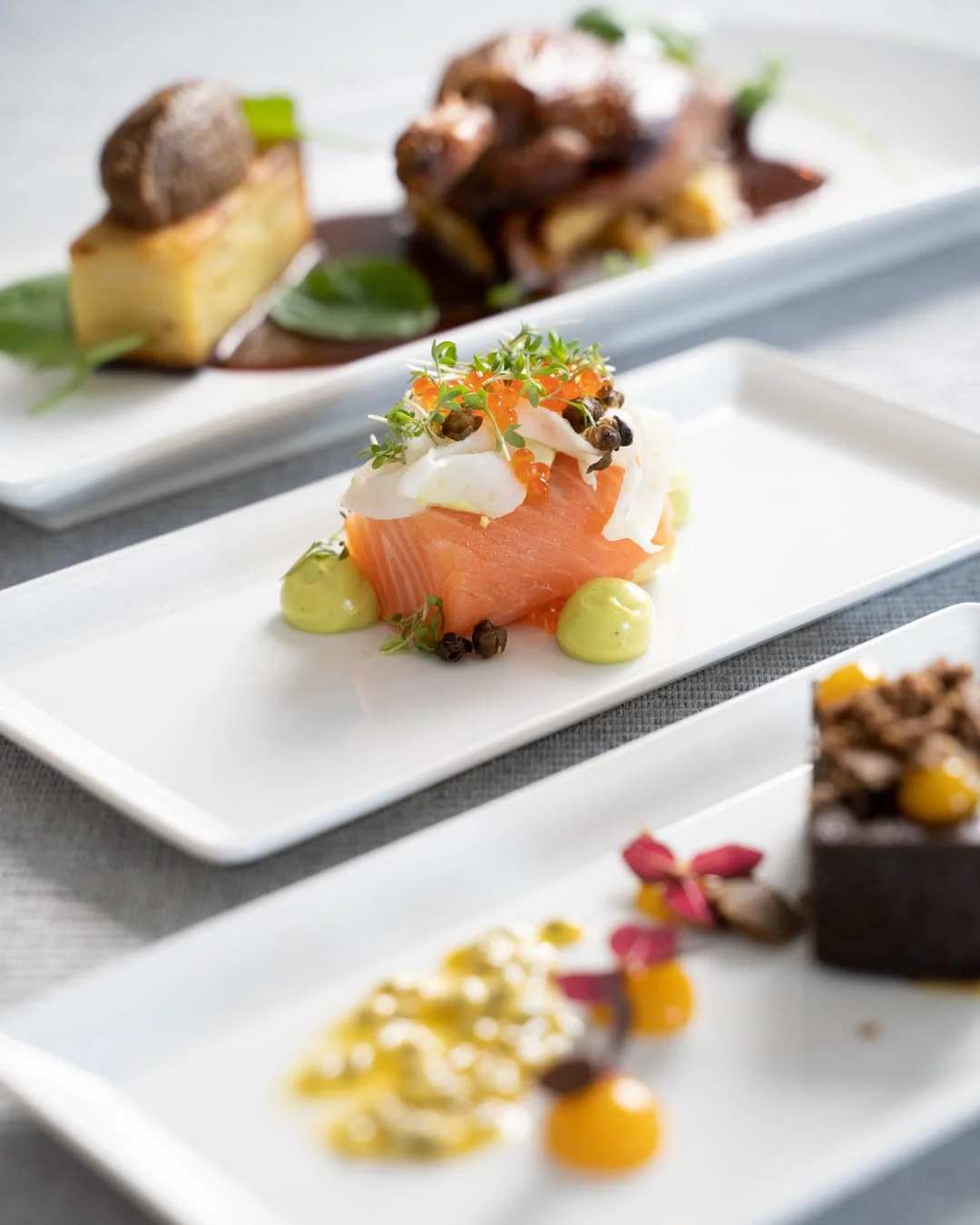 Eksklusiv fiskerett med laks, garnityr av små grønne urter, rømme, røkt laks og lakserogn på en hvit tallerken, med flere gourmetretter i bakgrunnen.