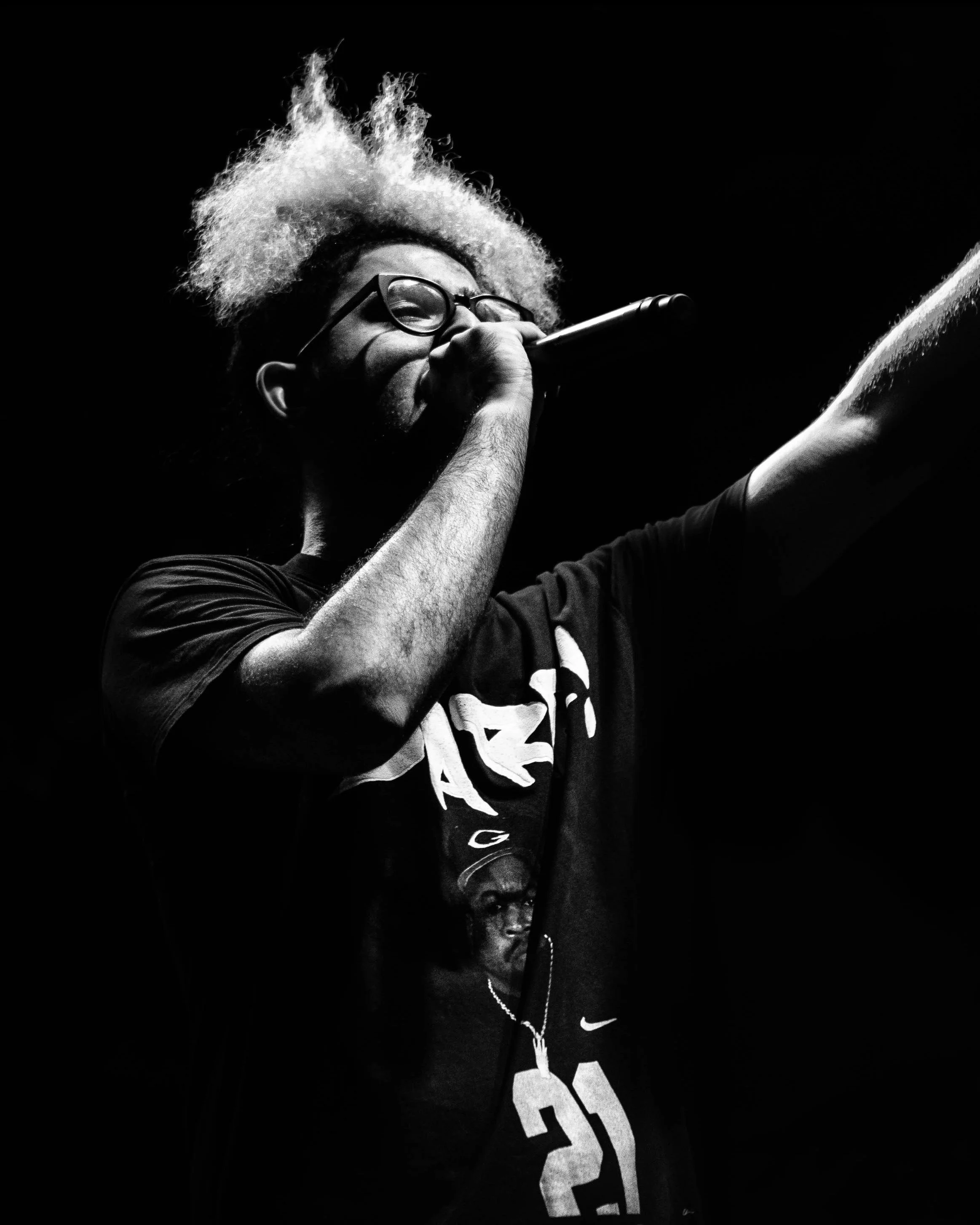 En mann med briller og blond hår som synger inn i en mikrofon på en mørk scene.