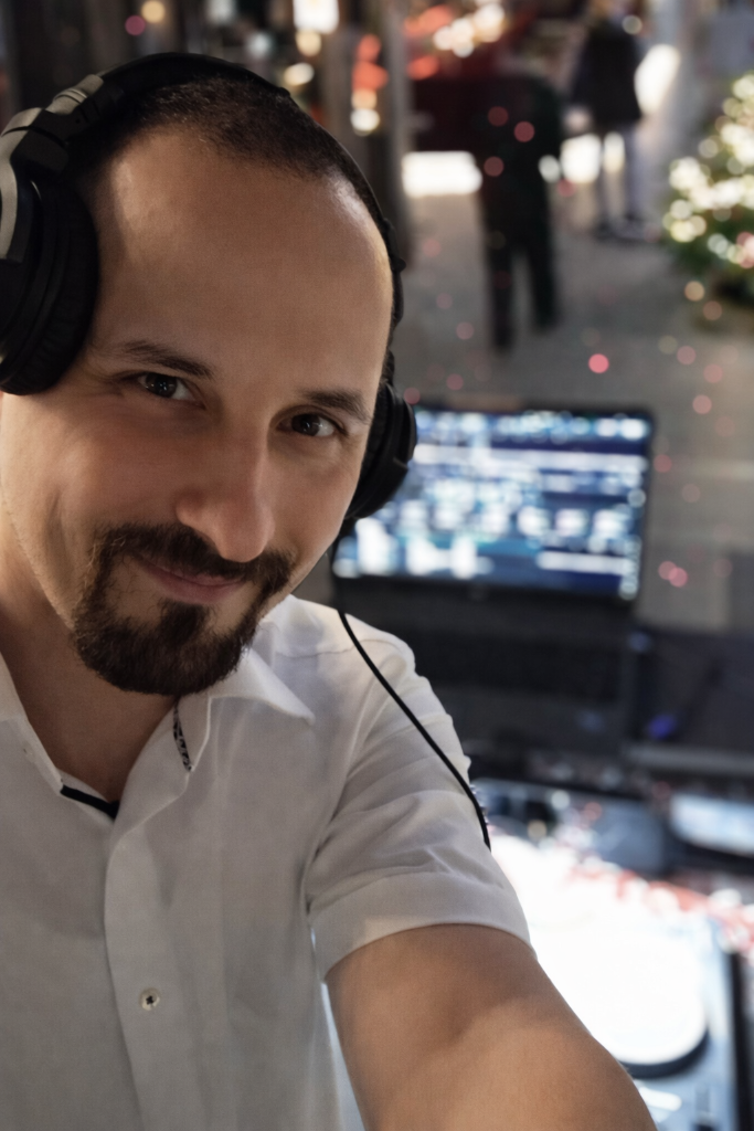 Ein Mann mit einem weißen Hemd trägt Kopfhörer und macht ein Selfie. Im Hintergrund ist ein Bildschirm mit einer Musik- oder DJ-Ausrüstung sichtbar, mit Lichter und Menschen in einer ungeklärten Umgebung.