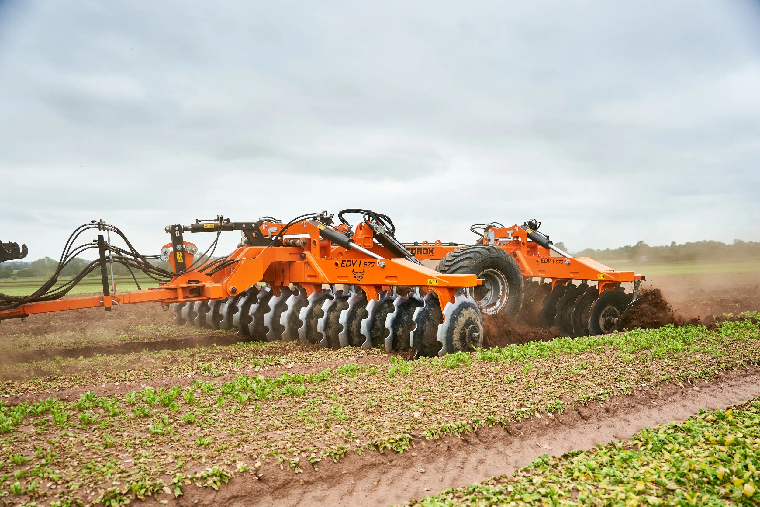 Agricultural tractor tilling a field with green crops during daytime under cloudy sky.