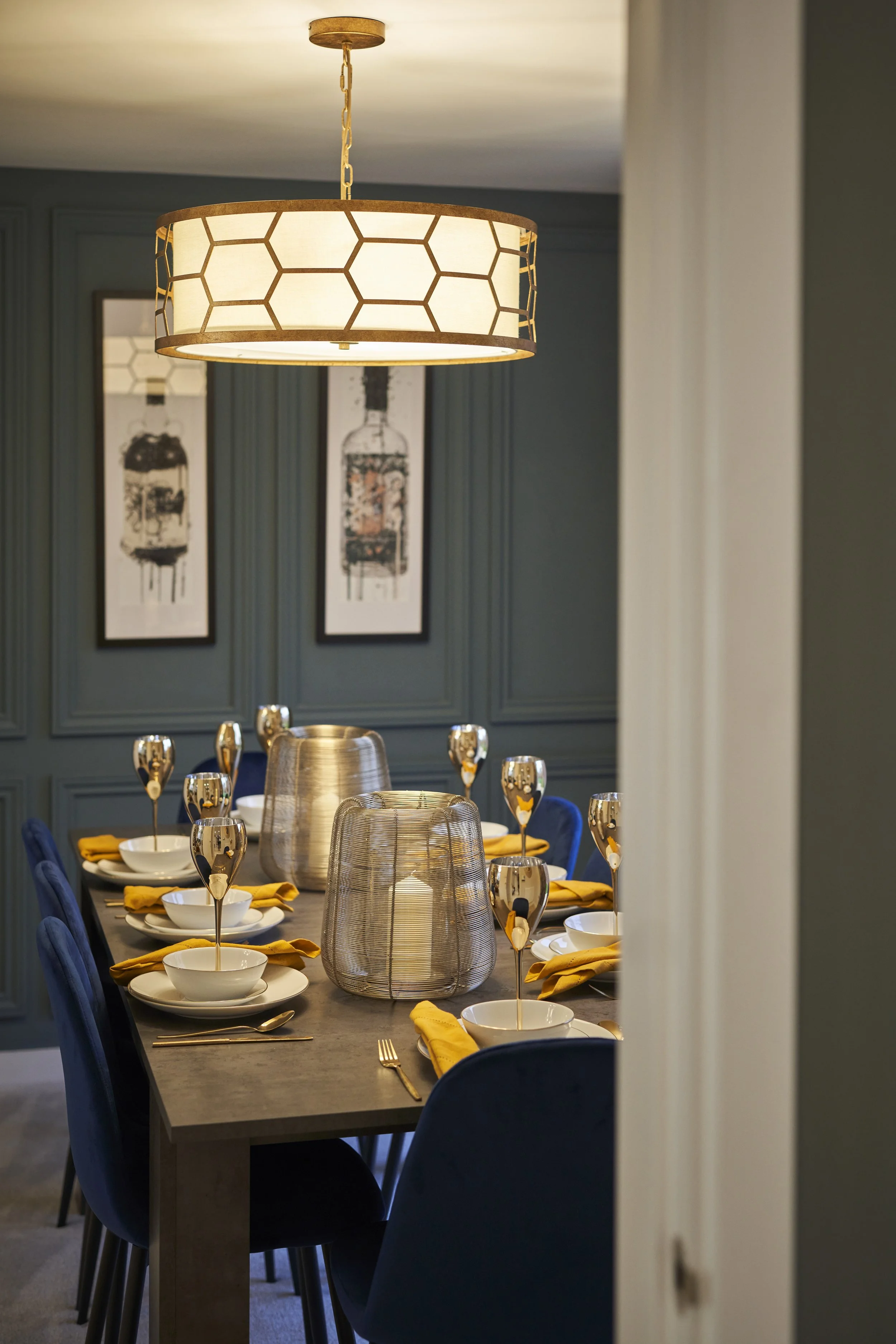 A dining room with a modern hanging light fixture, dark blue chairs, yellow napkins, white bowls, and metallic gold stemmed glasses set on a dark wooden table, with teal paneled walls and framed artwork in the background.