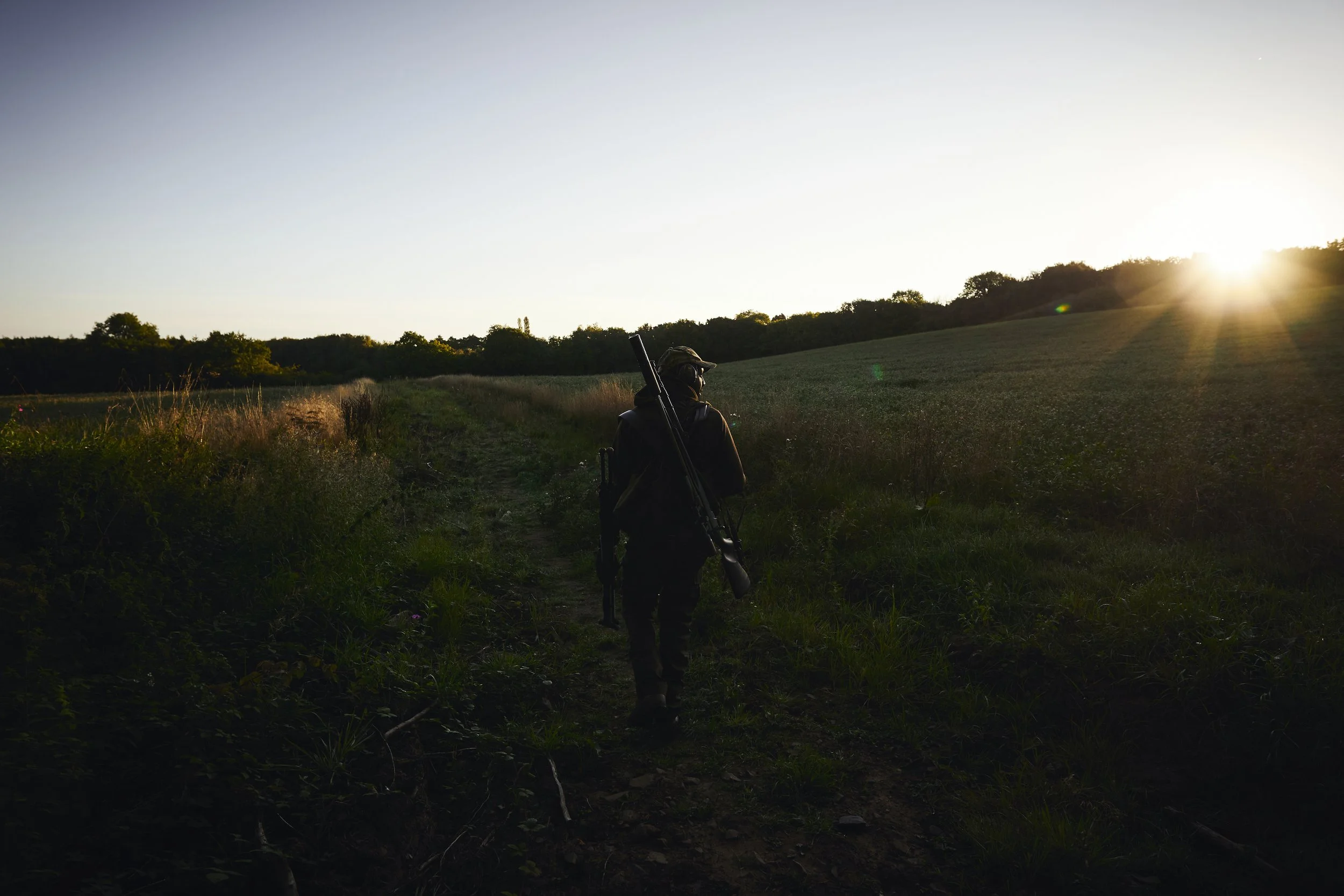 Person walking on a dirt path through a grassy field at sunset, carrying a rifle on their shoulder.