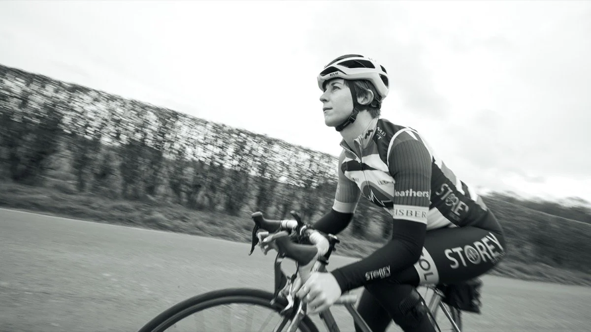A woman cycling outdoors on a road, wearing a helmet and cycling gear, with trees and cloudy sky in the background.