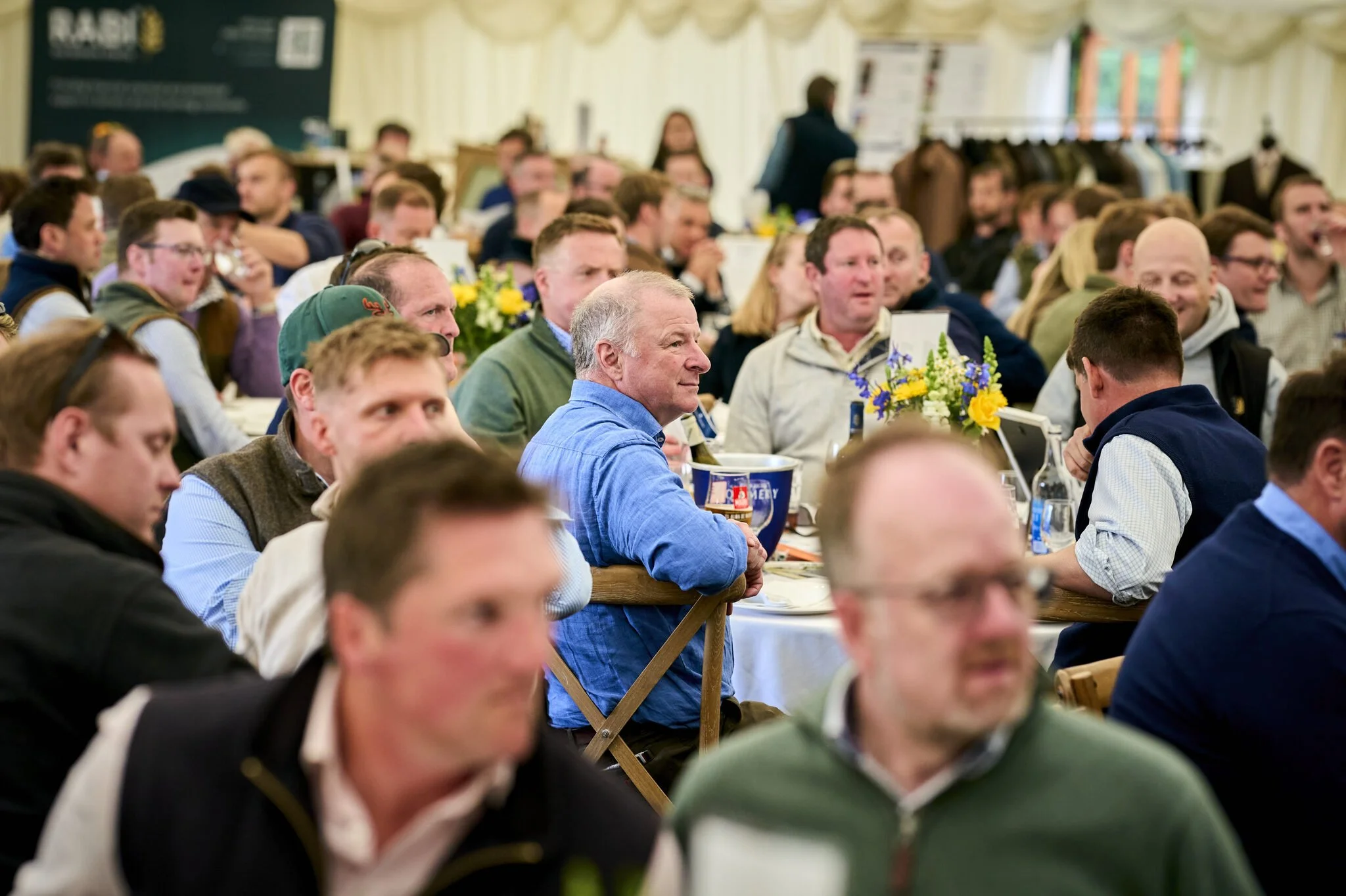 A large group of people seated at long tables in a tent, appearing to attend an event or gathering, with some people engaged in conversation and others listening.